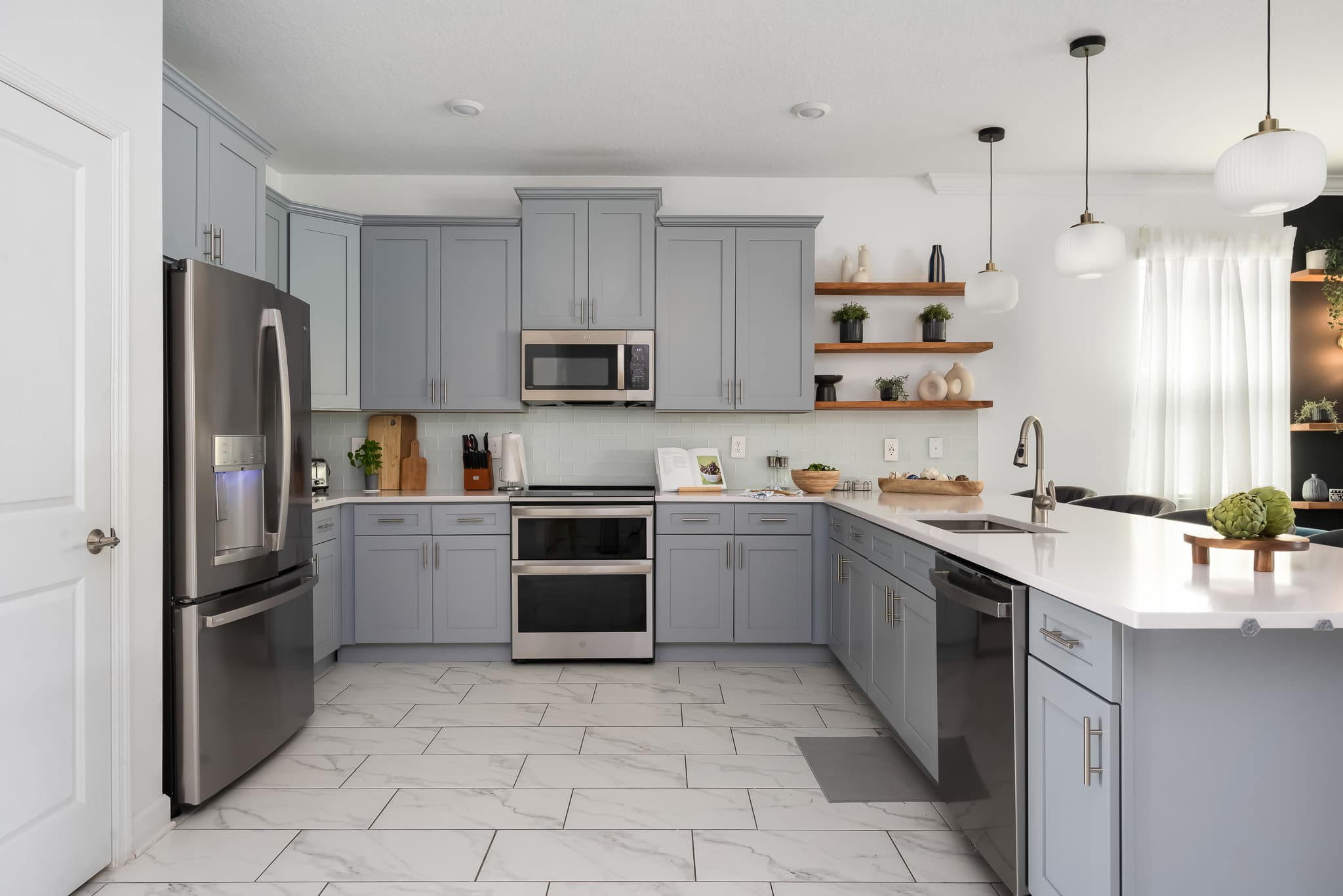 Wide shot of a modern kitchen featuring light gray shaker cabinets, white quartz countertops, stainless steel appliances including a double oven range, and a white subway tile backsplash.