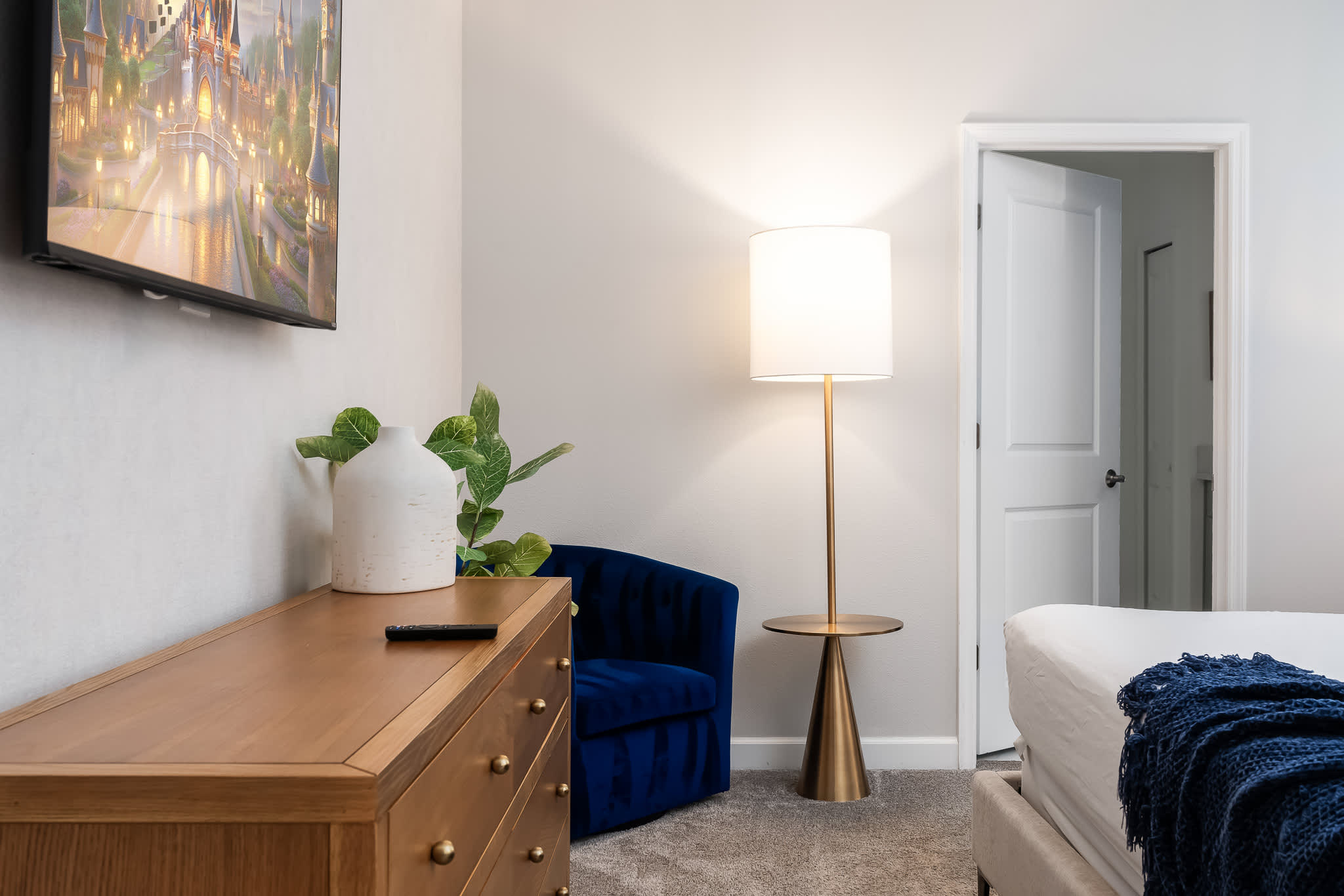 A corner of the bedroom shows a wooden dresser with a white vase and a faux plant, a wall-mounted TV displaying a castle, and a plush blue velvet accent chair. A modern gold floor lamp stands beside the chair, illuminating the space.