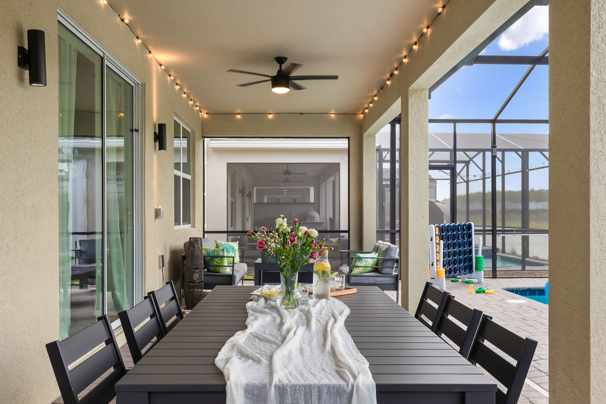 A covered outdoor patio area with string lights and a ceiling fan, featuring a long dark dining table set with a white runner and a floral centerpiece, and comfortable outdoor seating in the background.