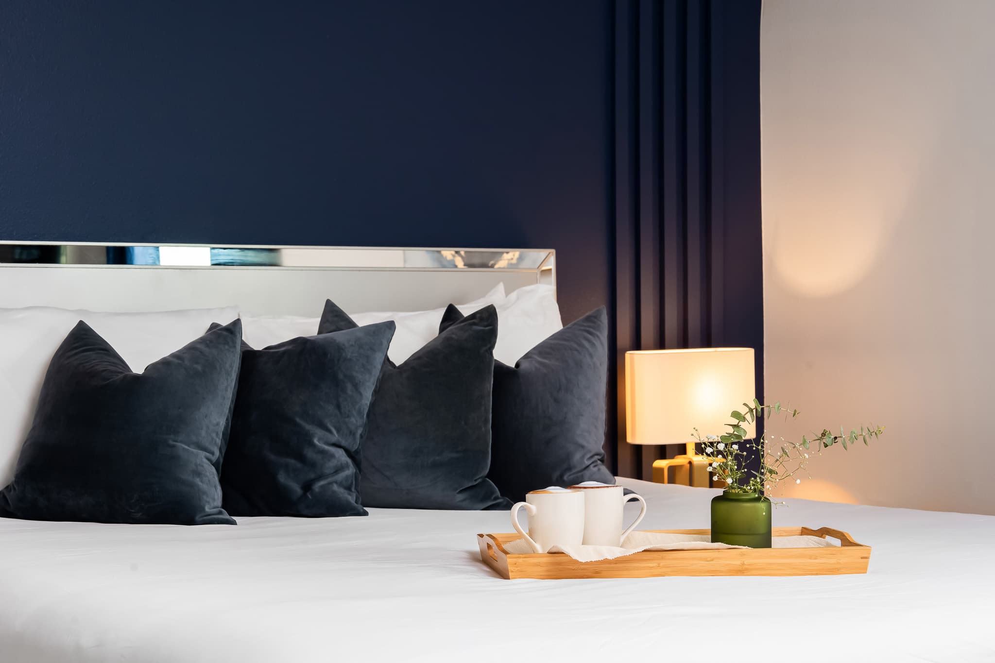 A close-up view of a bed with crisp white linens and dark blue velvet pillows, featuring a mirrored headboard and a wooden tray with two white mugs and a small green vase with foliage.