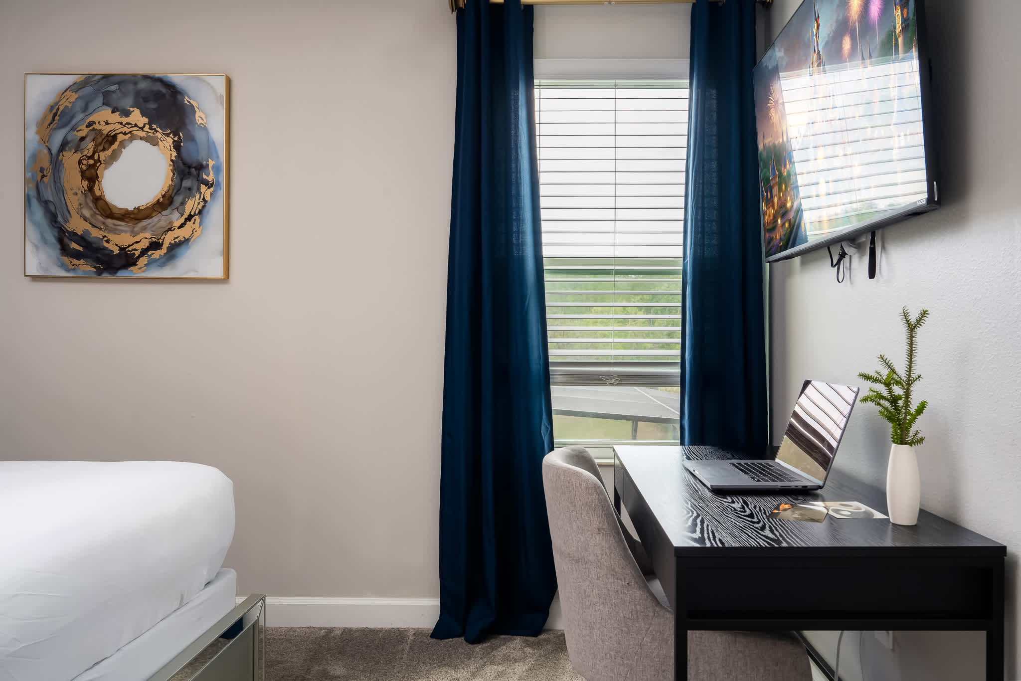 A bright corner of a bedroom featuring a black desk with a laptop and a white vase with greenery, positioned next to a window with blue curtains and blinds, and an abstract gold and blue artwork on the wall.