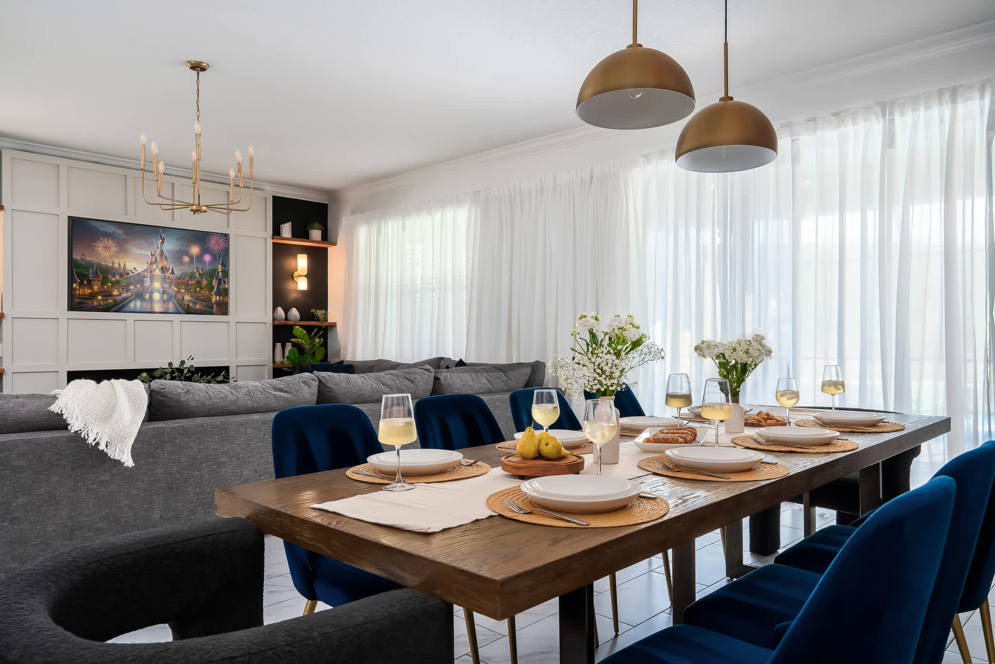Dining area with a rustic wooden table and blue velvet chairs, offering a view into the adjacent living room with its gray sectional and entertainment center. The space is bright with white sheer curtains.