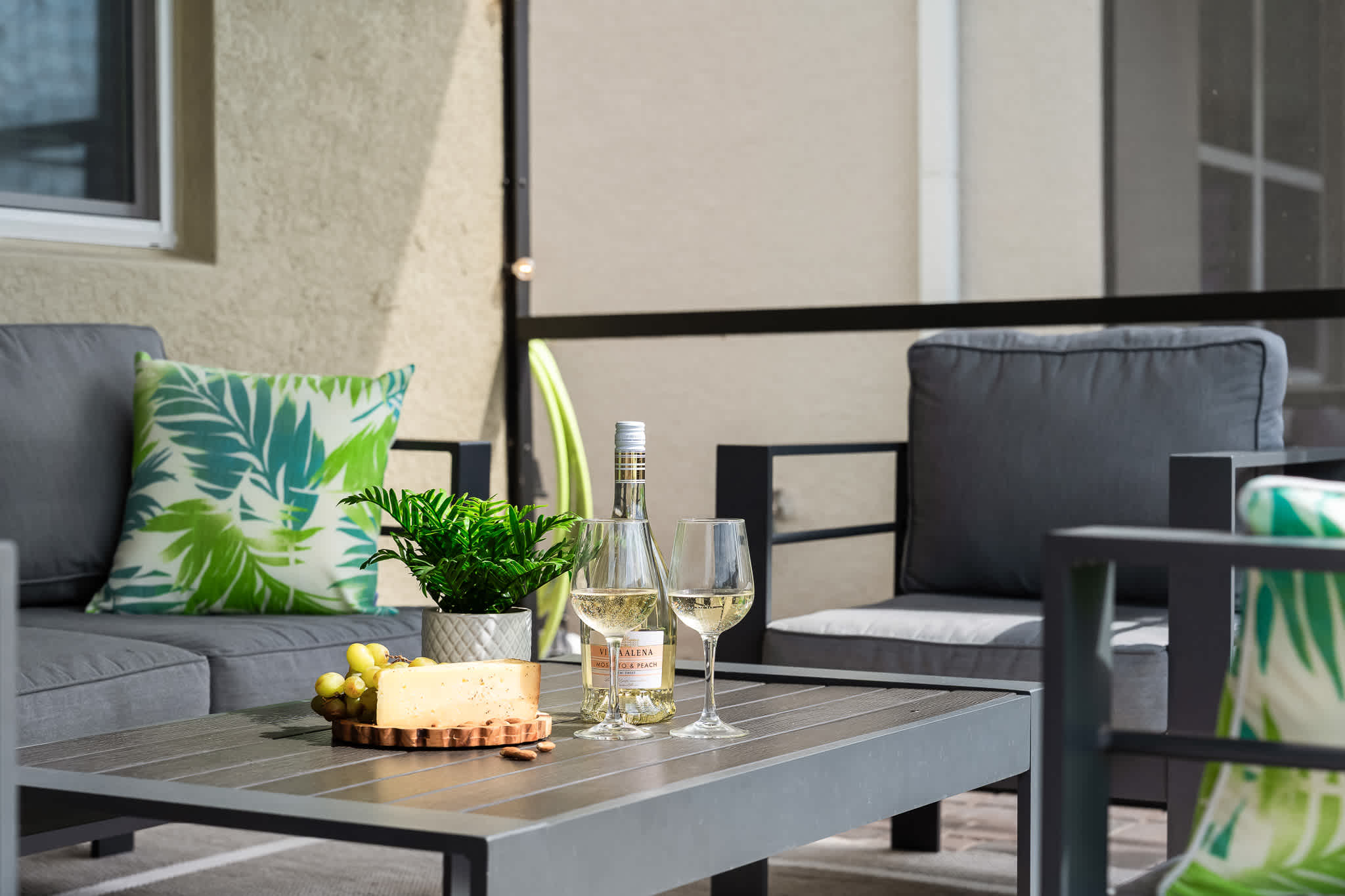 A bottle of white wine, two wine glasses, a cheese board with cheese and grapes, and a potted plant are arranged on a dark grey outdoor patio table, with grey outdoor seating in the background.