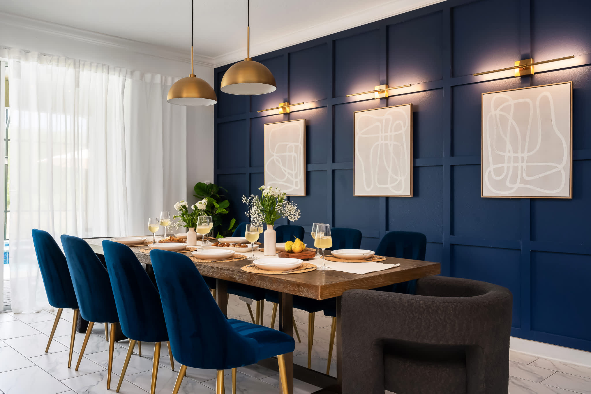 Elegant dining area featuring a long wooden table set for eight with blue velvet chairs, illuminated by two gold pendant lights. A dark blue paneled accent wall with sconces and abstract art adds sophistication.