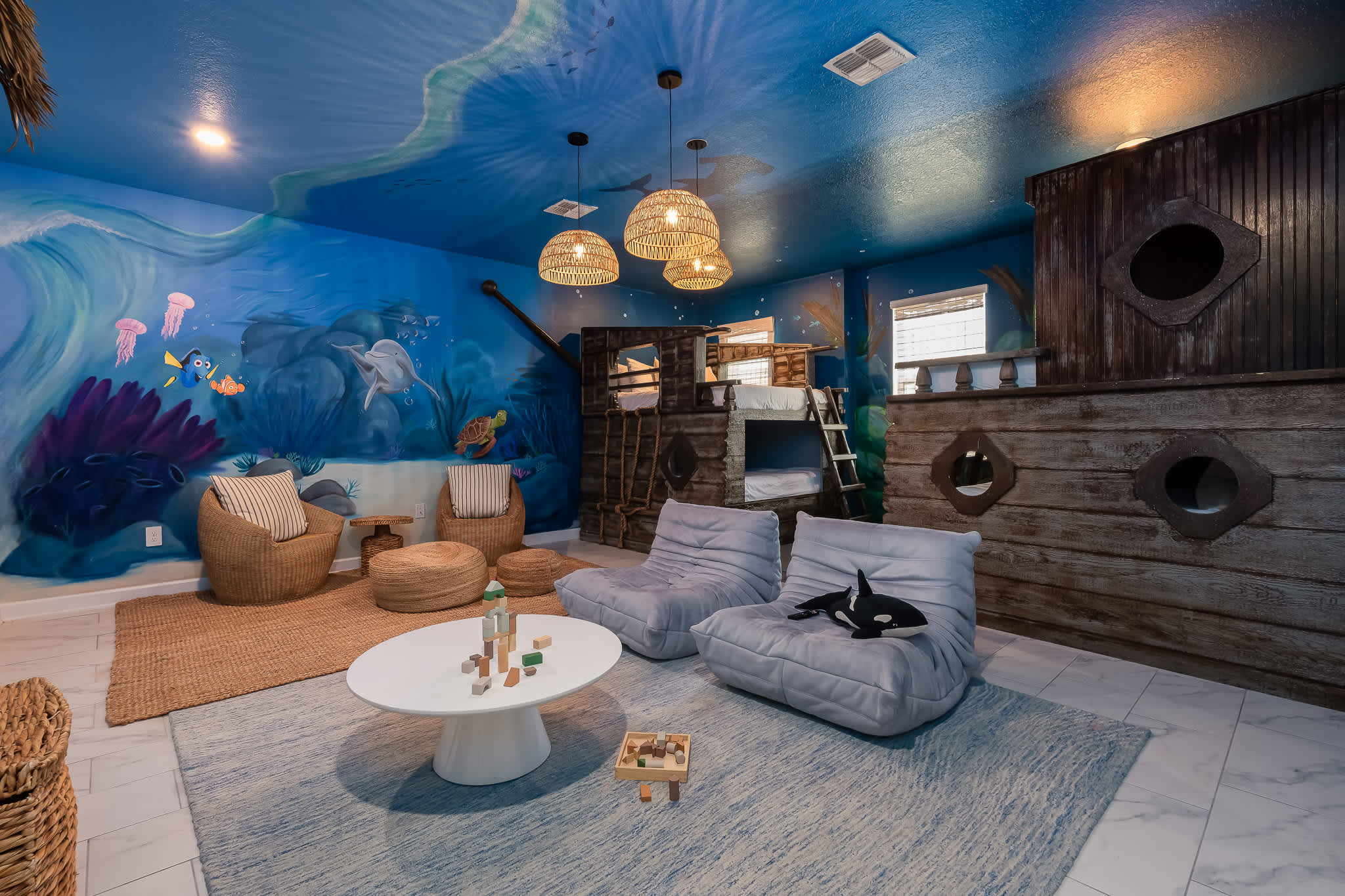Vibrant underwater-themed bedroom with blue walls and ceiling murals, featuring a large wooden ship-themed bunk bed and a separate seating area with wicker chairs and a white coffee table.