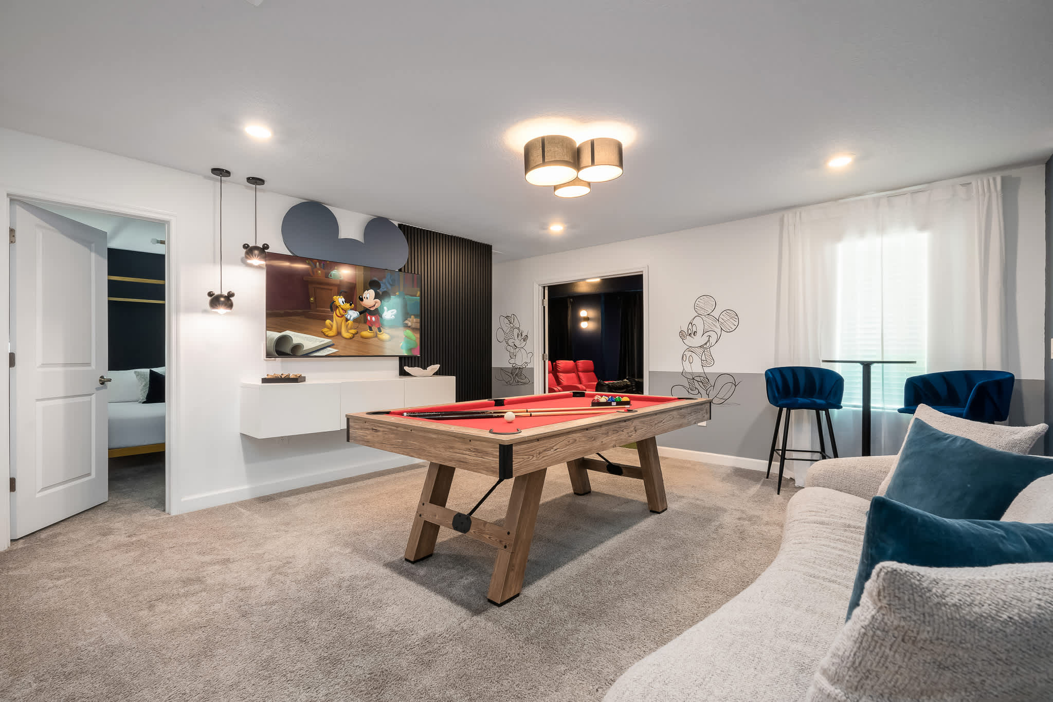 A vibrant game room features a red-felted pool table at its center, a large TV with Mickey Mouse ears decor, and Mickey Mouse wall art, with a glimpse of a dark media room beyond an open doorway.