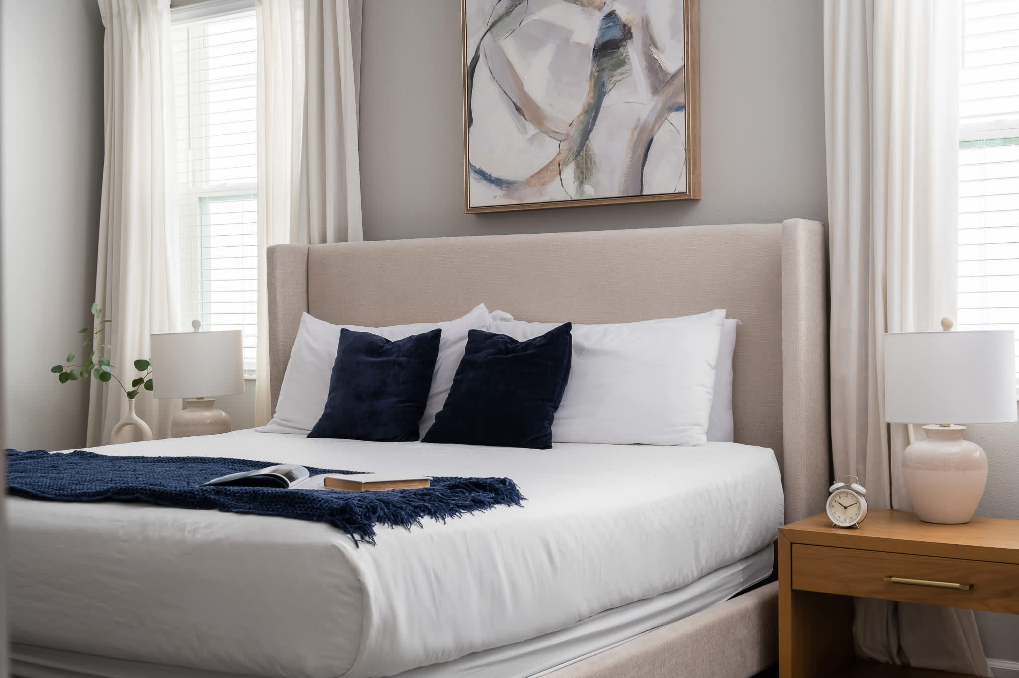 A close-up of a king-sized bed with a tall, light gray upholstered headboard, crisp white bedding, and two navy velvet throw pillows. Light wood nightstands flank the bed, each with a ceramic lamp, and a blue throw blanket is draped across the foot of the bed.