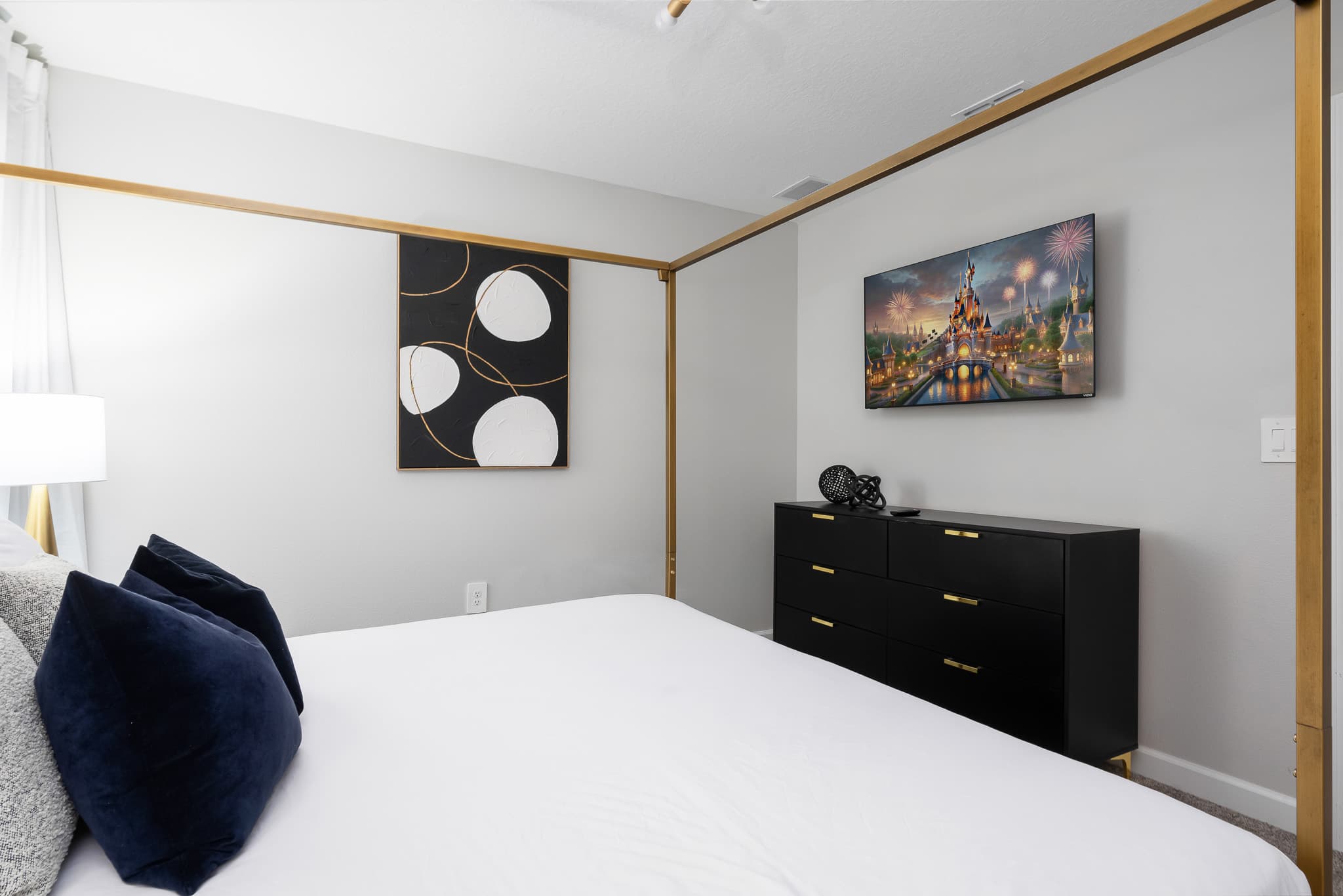 View of the elegant bedroom showing a wall-mounted TV displaying a fireworks scene over a castle, a black dresser with gold accents, and a gold four-poster king bed with white bedding.