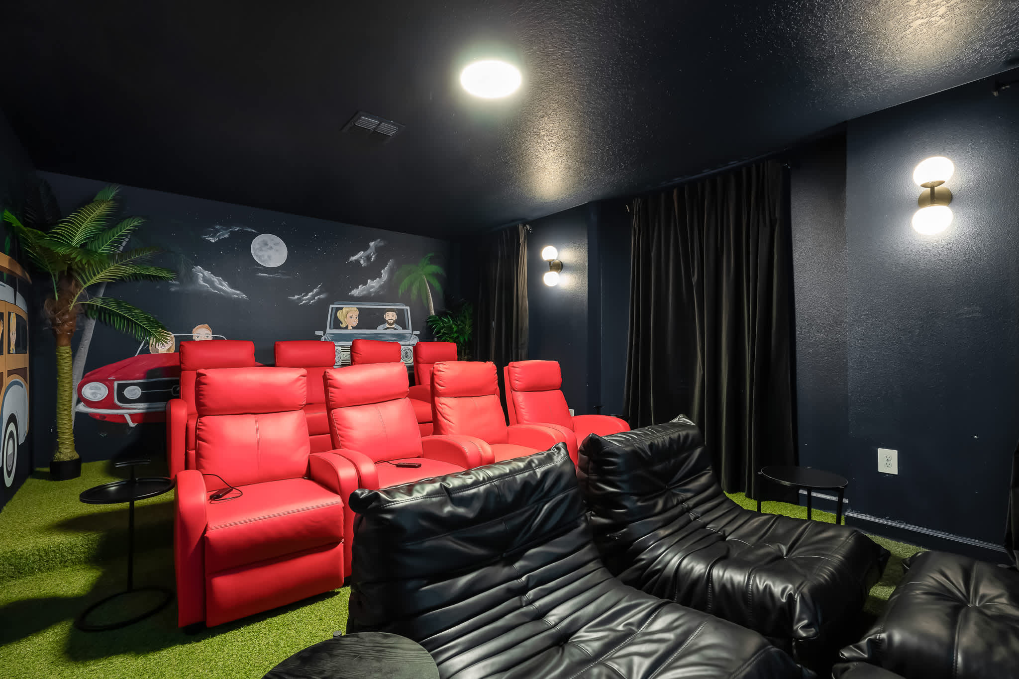 A dedicated media room features rows of comfortable red recliners and black bean bag chairs on a green turf-like floor, with dark walls adorned with a car-themed mural and palm trees.