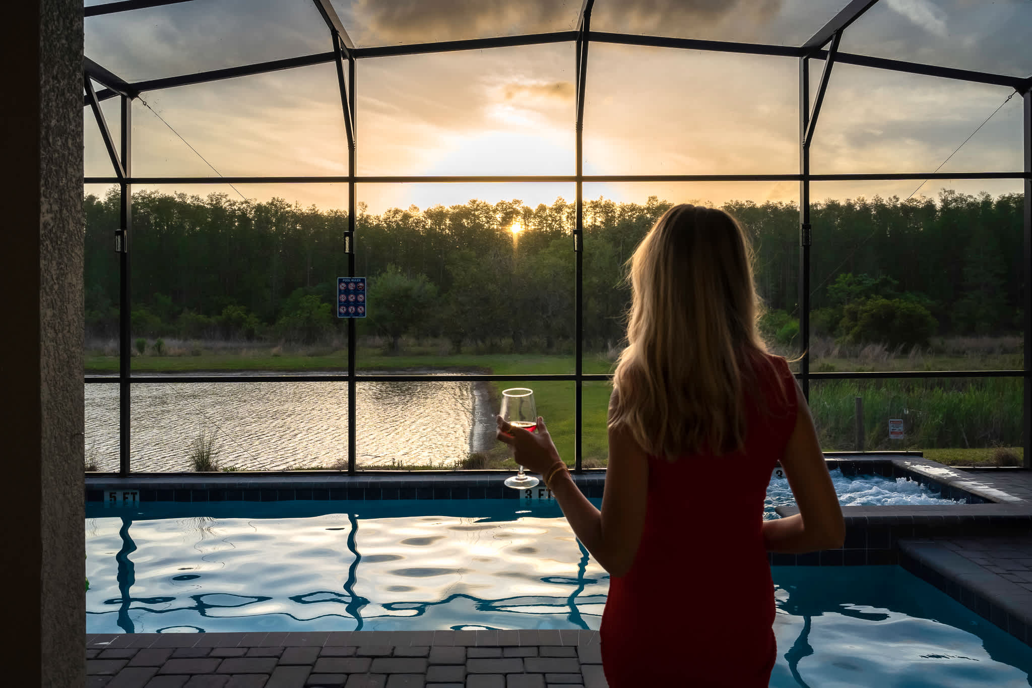 A woman in a red dress stands by the private pool, holding a glass of wine, admiring the vibrant sunset over a tranquil lake and forest from the screened patio.