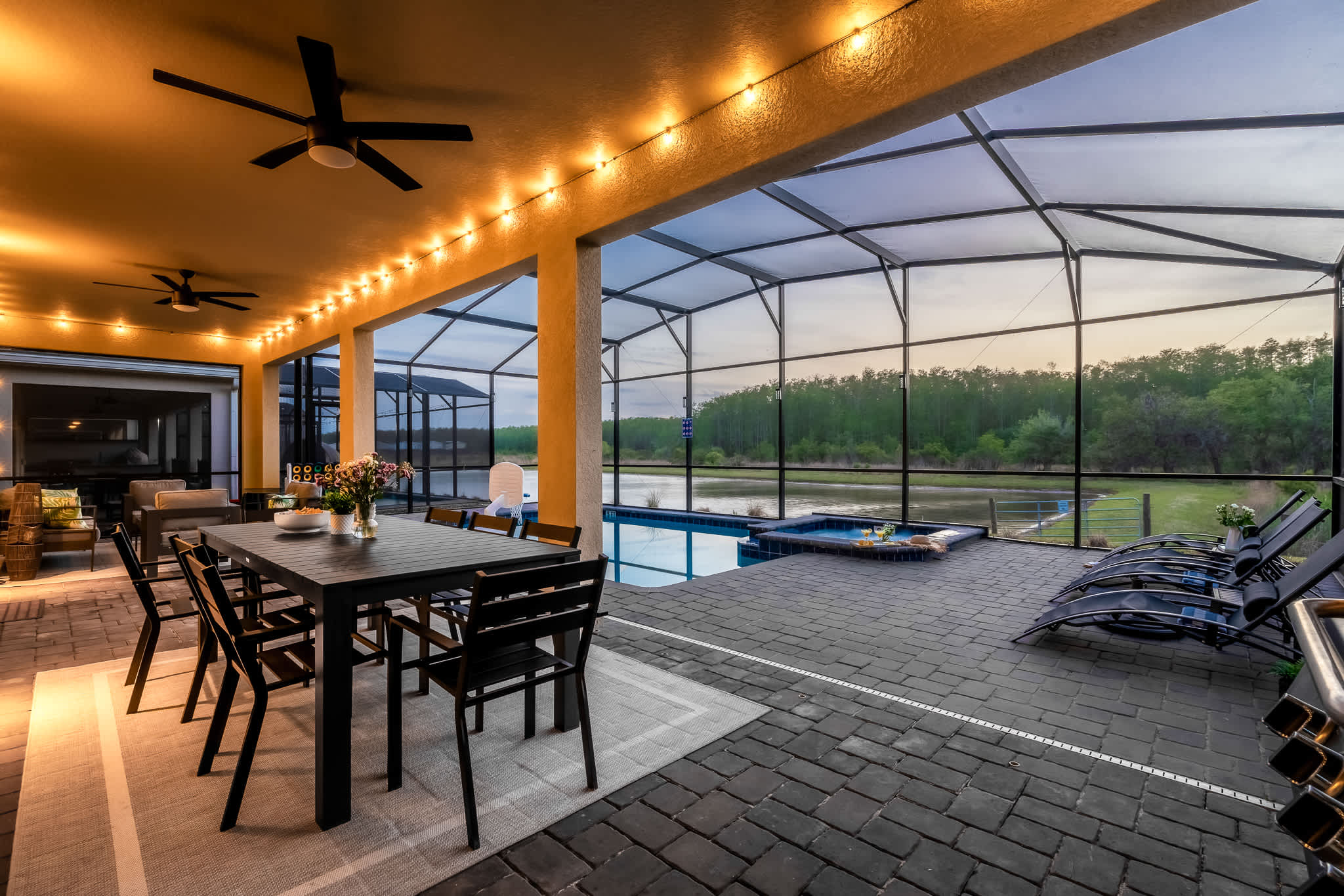 Spacious screened-in outdoor patio with a large dark dining table, several lounge chairs, a private pool, and hot tub, illuminated by string lights at dusk.