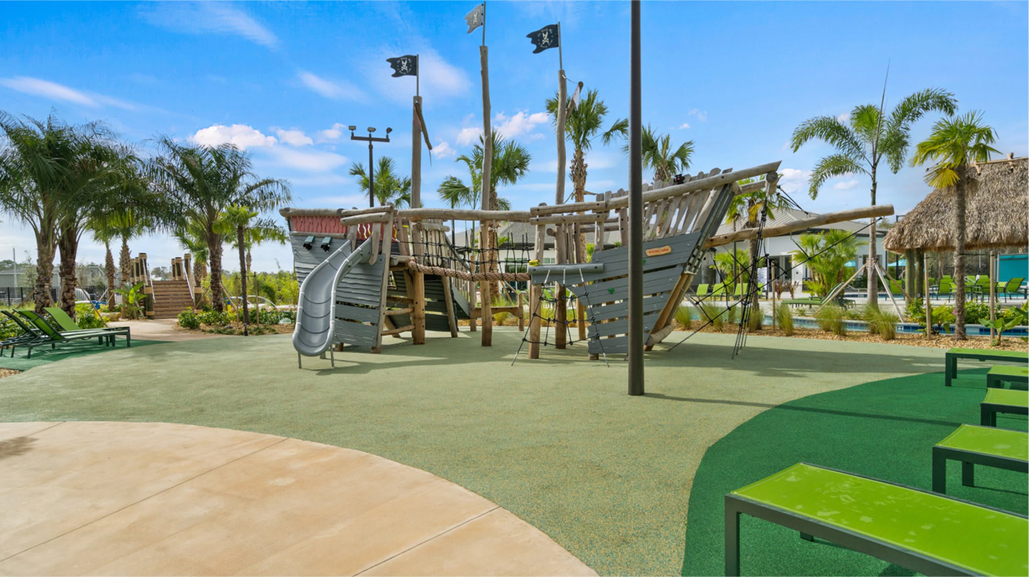 Creative children's playground featuring a large wooden pirate ship structure with slides, climbing nets, and pirate flags, set on a green soft-fall surface, surrounded by palm trees and lounge seating.