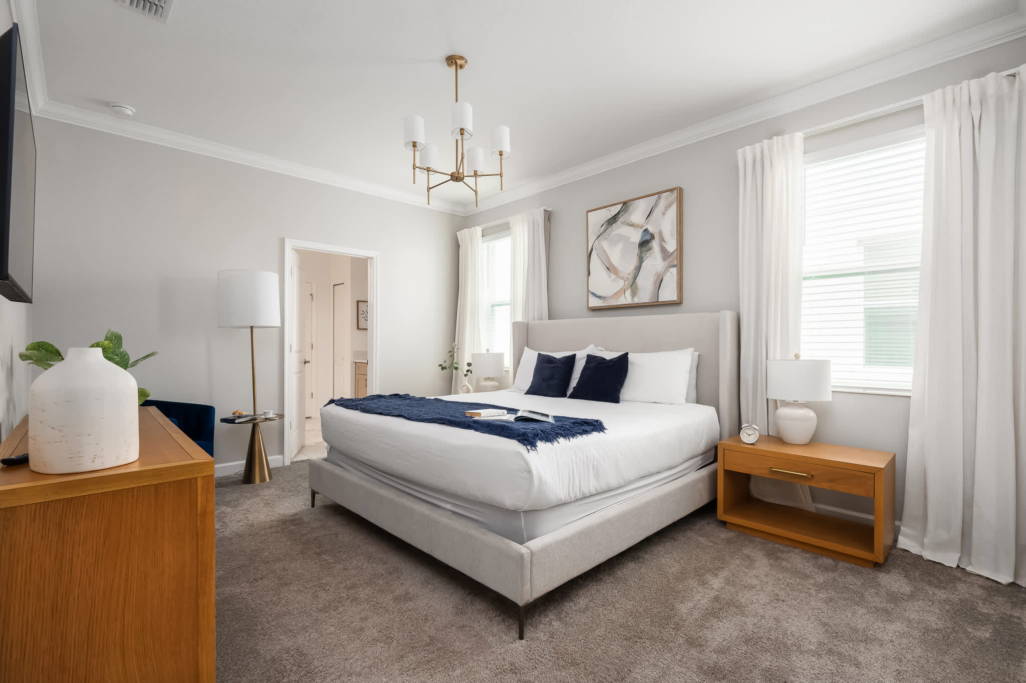 A spacious bedroom features a king-sized bed with a light gray upholstered headboard, a blue throw blanket, and navy pillows, complemented by light wood nightstands and a dresser. A gold chandelier hangs from the ceiling, and a TV displays a castle scene.