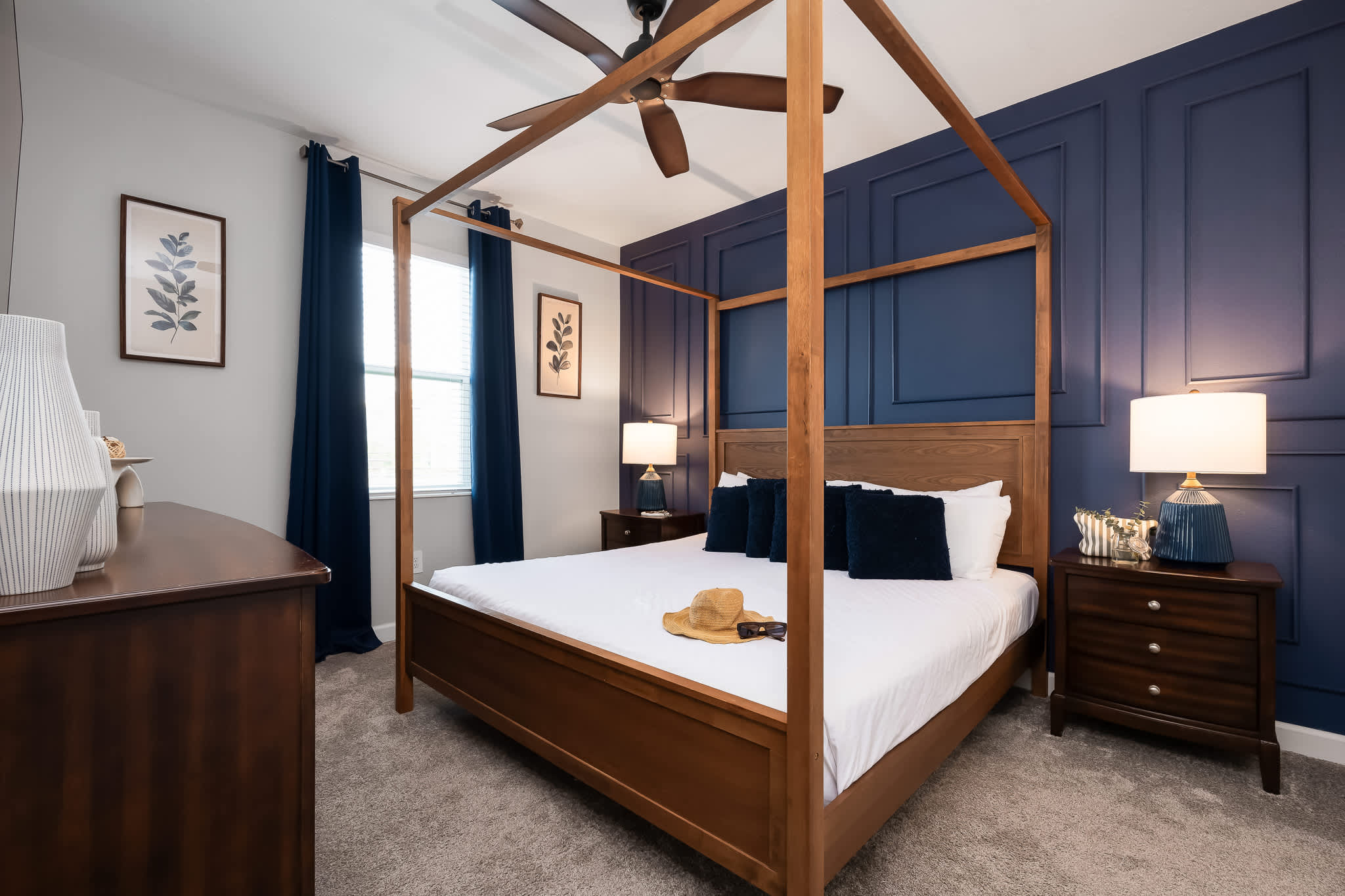 Spacious bedroom with a wooden four-poster king bed, dark blue accent wall with wainscoting, dark wood nightstands, and a ceiling fan.