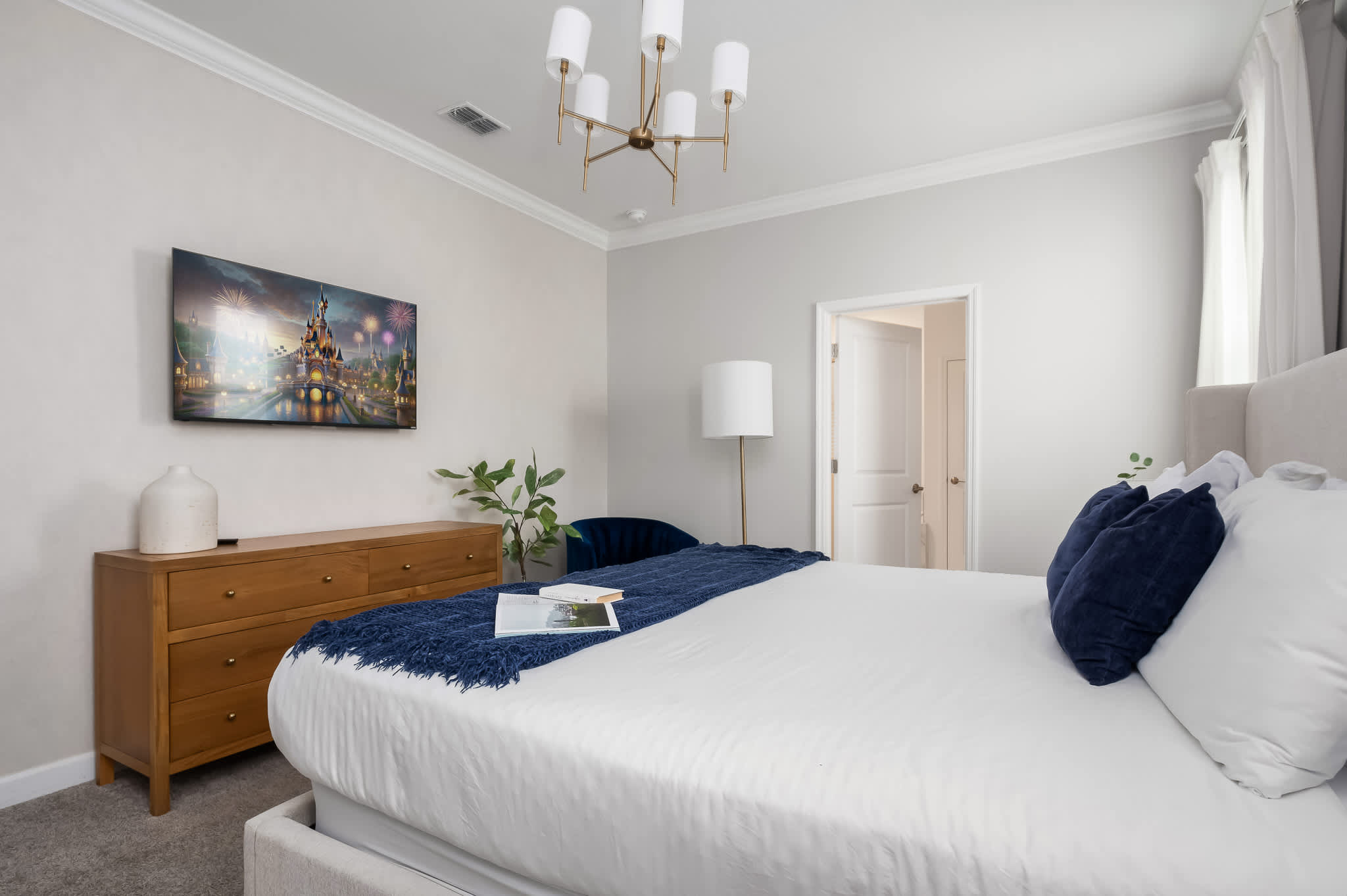 This bright bedroom features a king-sized bed with a light gray headboard, a blue throw, and navy pillows, facing a wooden dresser with a TV mounted above it. A modern gold floor lamp and a blue velvet accent chair are visible near the doorway.