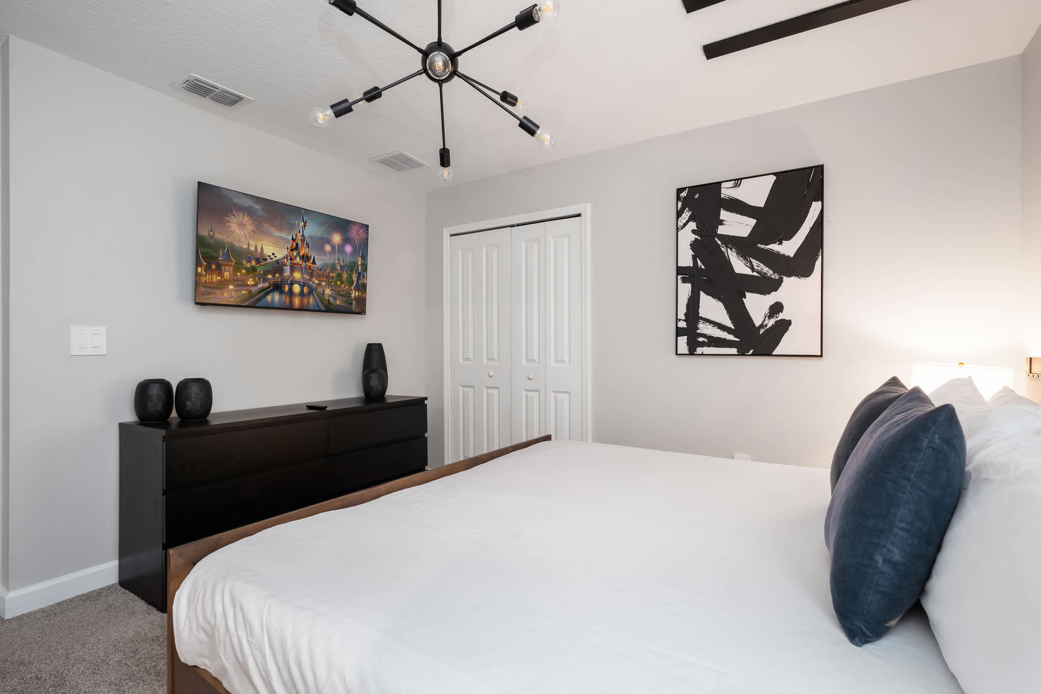 Modern bedroom with a king-sized bed, a black dresser with a wall-mounted TV, and a striking black and white abstract painting, complemented by a unique sputnik chandelier.