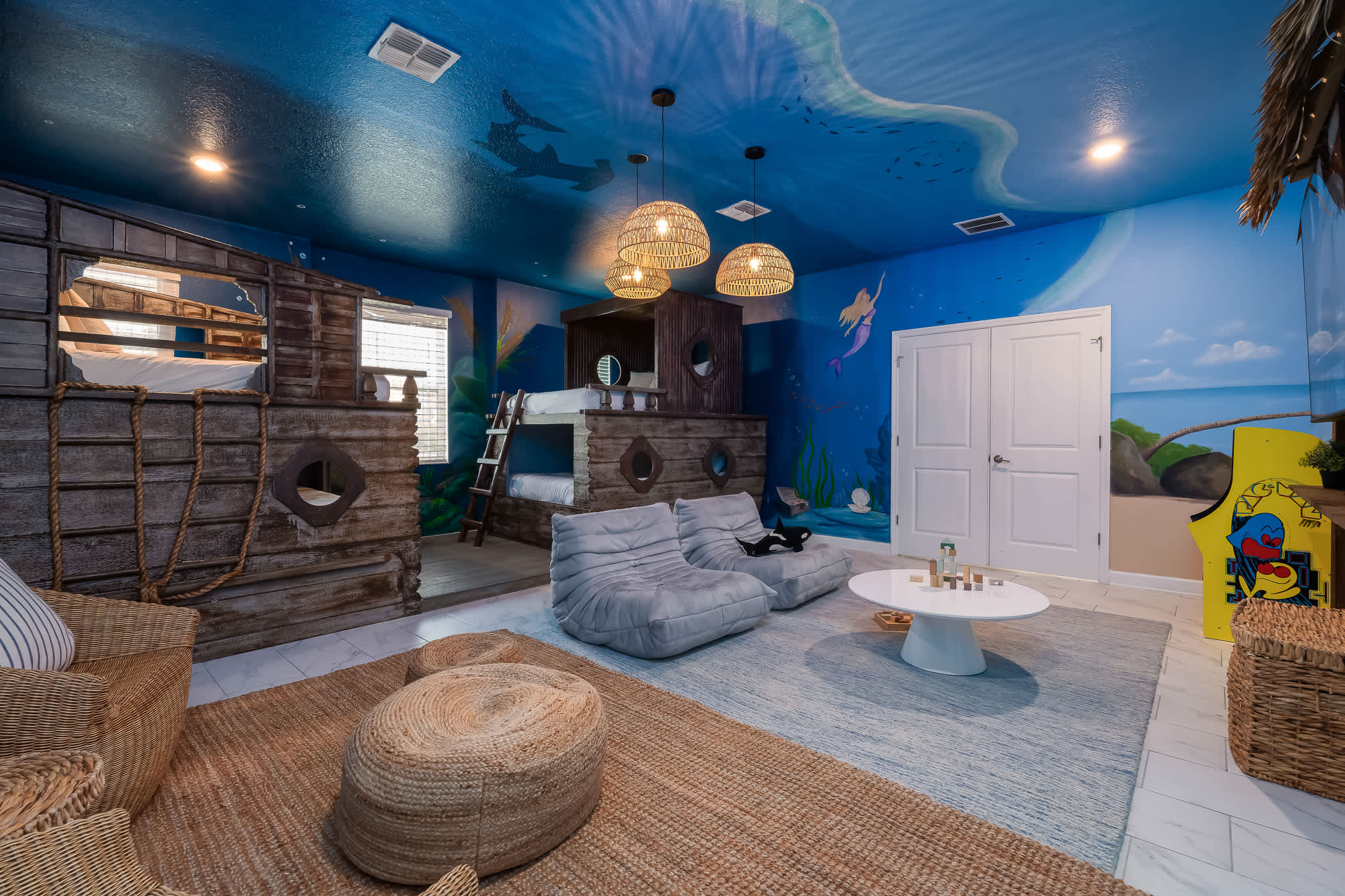 A vibrant, ocean-themed room features a blue ceiling painted with underwater scenes, two rustic wooden bunk bed structures, and a central play area with two gray bean bag chairs and a white coffee table. Woven pendant lights hang from the ceiling, adding to the playful ambiance.