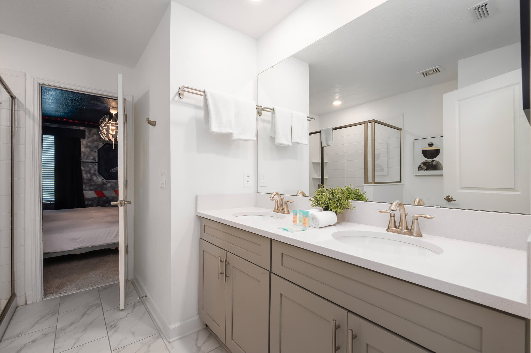 Modern bathroom with a double vanity featuring light gray cabinets, white countertops, and dual sinks with brushed nickel faucets, a large mirror, and white towels, with a glimpse into the themed bedroom.