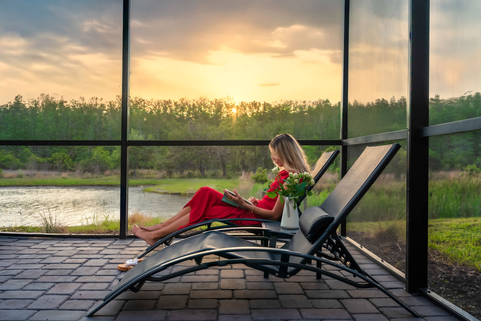 A woman in a red dress relaxes on a dark lounge chair on a screened-in paver patio, reading a book with a vase of flowers beside her, overlooking a lake at sunset.