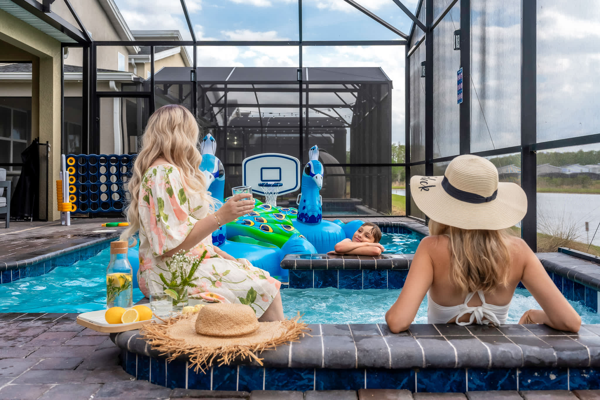 An outdoor screened-in pool area with two women and a child enjoying the water, featuring large inflatable peacock floats, a basketball hoop, and a view of a lake and trees in the background.