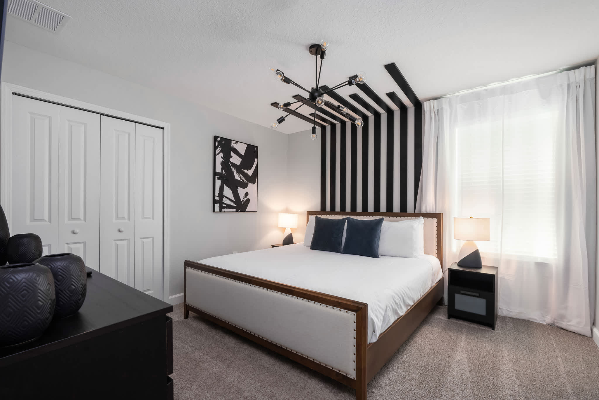 Stylish bedroom featuring a king-sized bed with a white upholstered headboard, black and white striped accent wall, a modern sputnik chandelier, and a large abstract artwork.