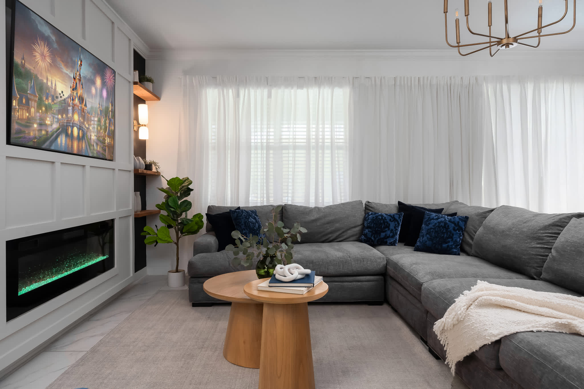 Bright living room featuring a large gray sectional sofa with blue accent pillows, facing a white paneled entertainment wall with a mounted TV and an electric fireplace. Sheer white curtains cover large windows.