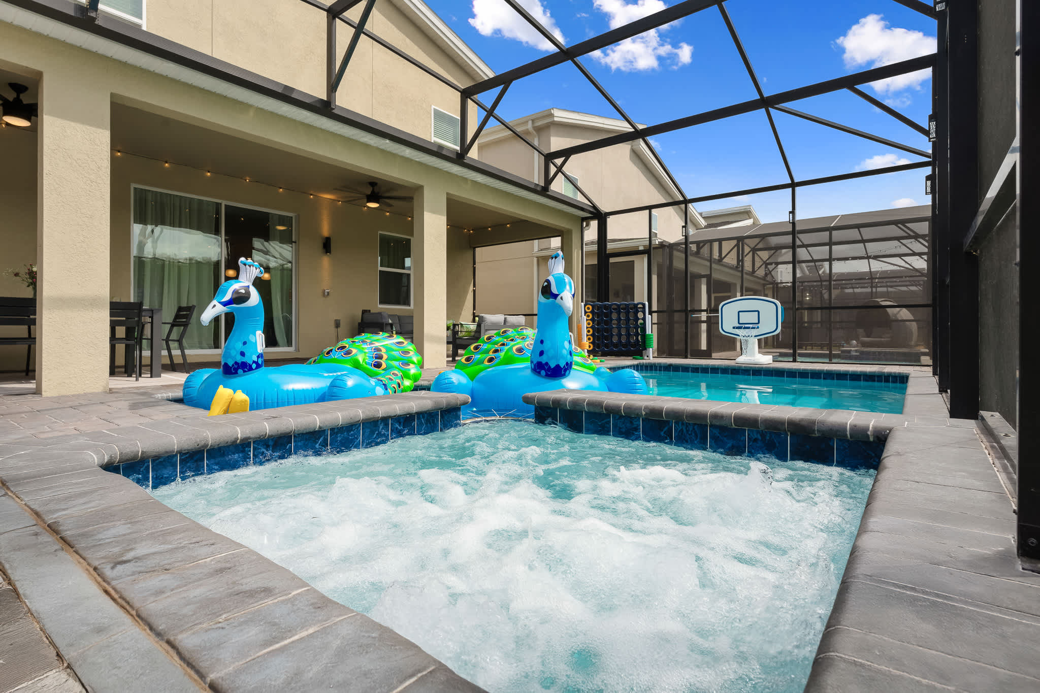A bubbling hot tub is featured in the foreground of a screened-in pool area, with large peacock-shaped pool floats in the adjacent main pool. The paved deck and surrounding enclosure are visible, with a house exterior in the background.