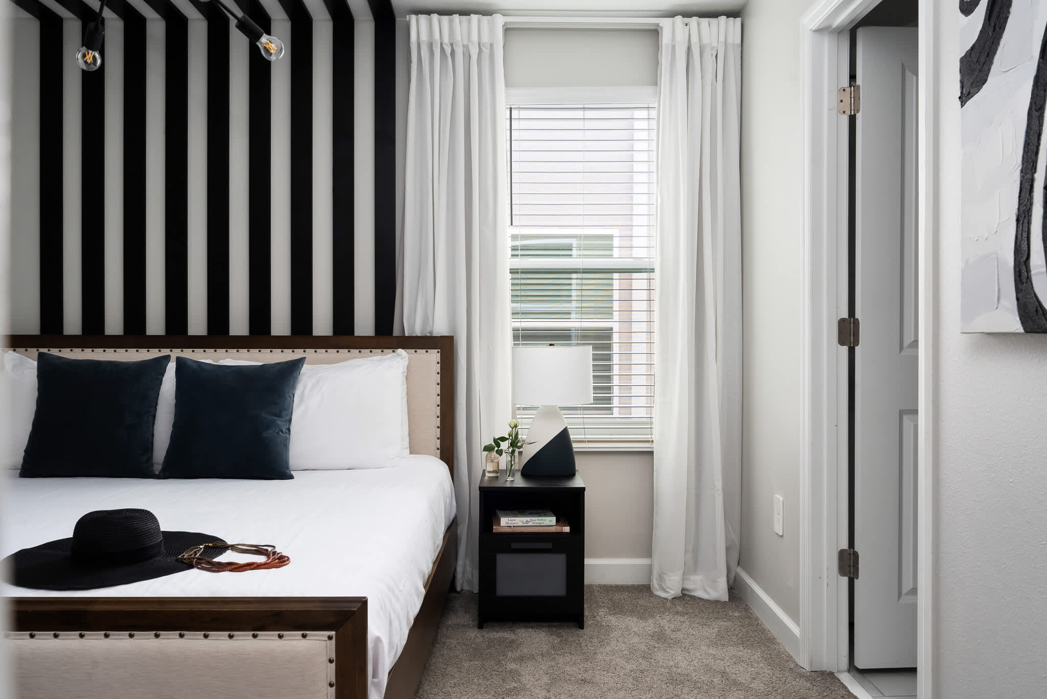 Contemporary bedroom with a king-sized bed, a black nightstand with a lamp, a window with white blinds and curtains, and a bold black and white striped accent wall behind the bed.