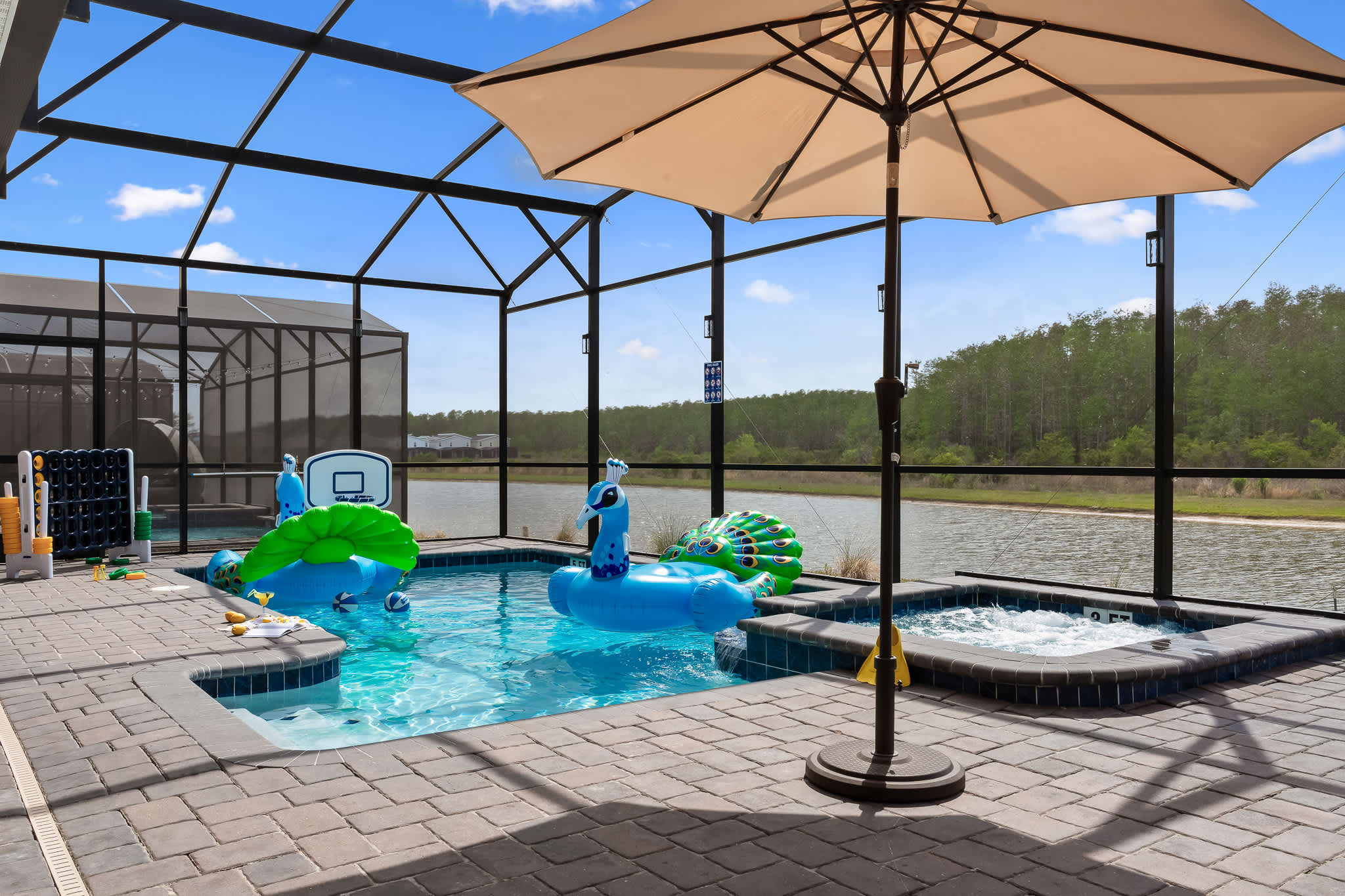 A spacious screened-in pool and hot tub area with brick pavers, featuring several inflatable peacock floats in the pool, a large patio umbrella, and a serene view of a lake and lush trees beyond the enclosure.