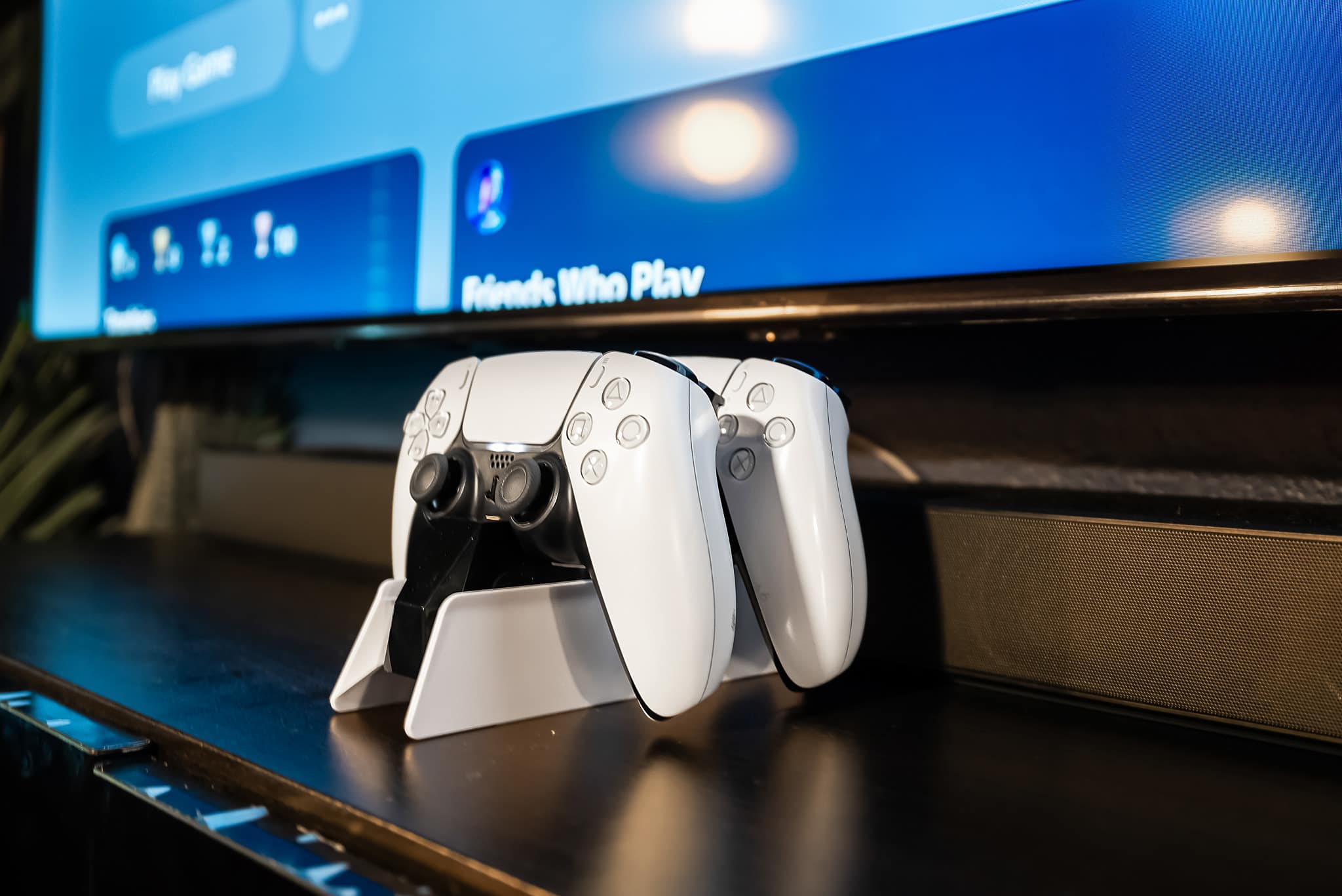 Two white PlayStation 5 controllers are neatly placed on a charging dock in front of a large TV screen displaying a game console interface, suggesting a dedicated gaming setup.