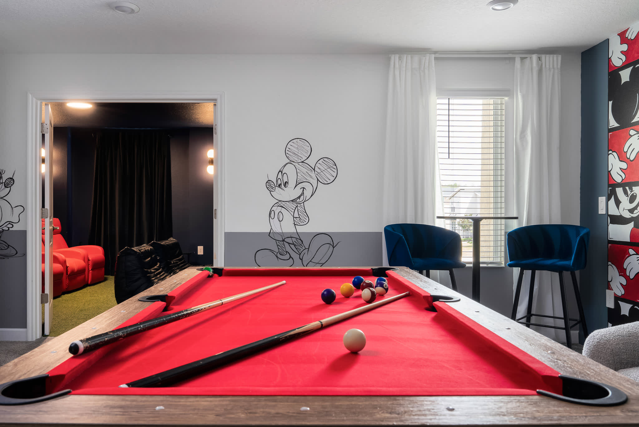 A close-up view of a red-felted pool table with cues and balls, set in a game room featuring Mickey Mouse wall art and two blue bar stools next to a window.