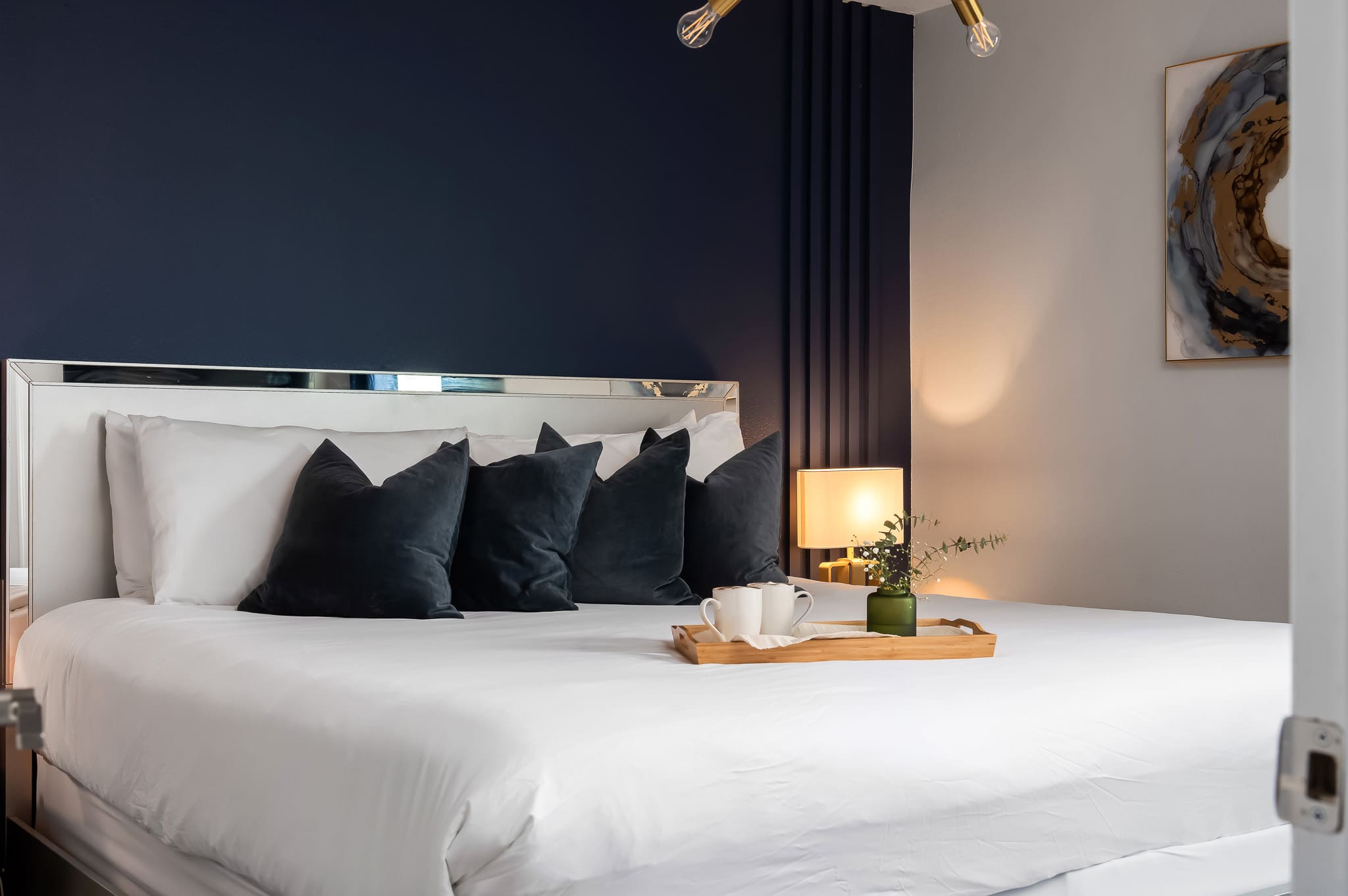 A modern bedroom with a dark blue accent wall behind a white bed with a mirrored headboard, adorned with white and dark blue velvet pillows, and a wooden tray with mugs and a plant on the bed.