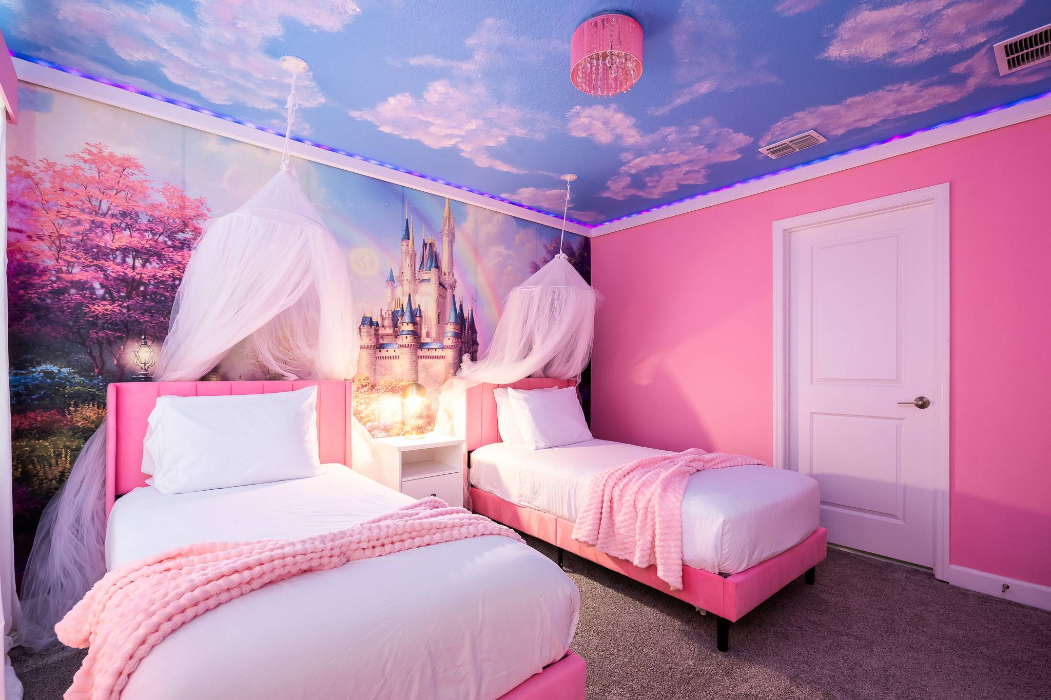 Whimsical princess-themed bedroom with two pink velvet twin beds, white canopies, and a vibrant wall mural depicting a castle and a magical forest under a cloud-painted ceiling with LED lighting.