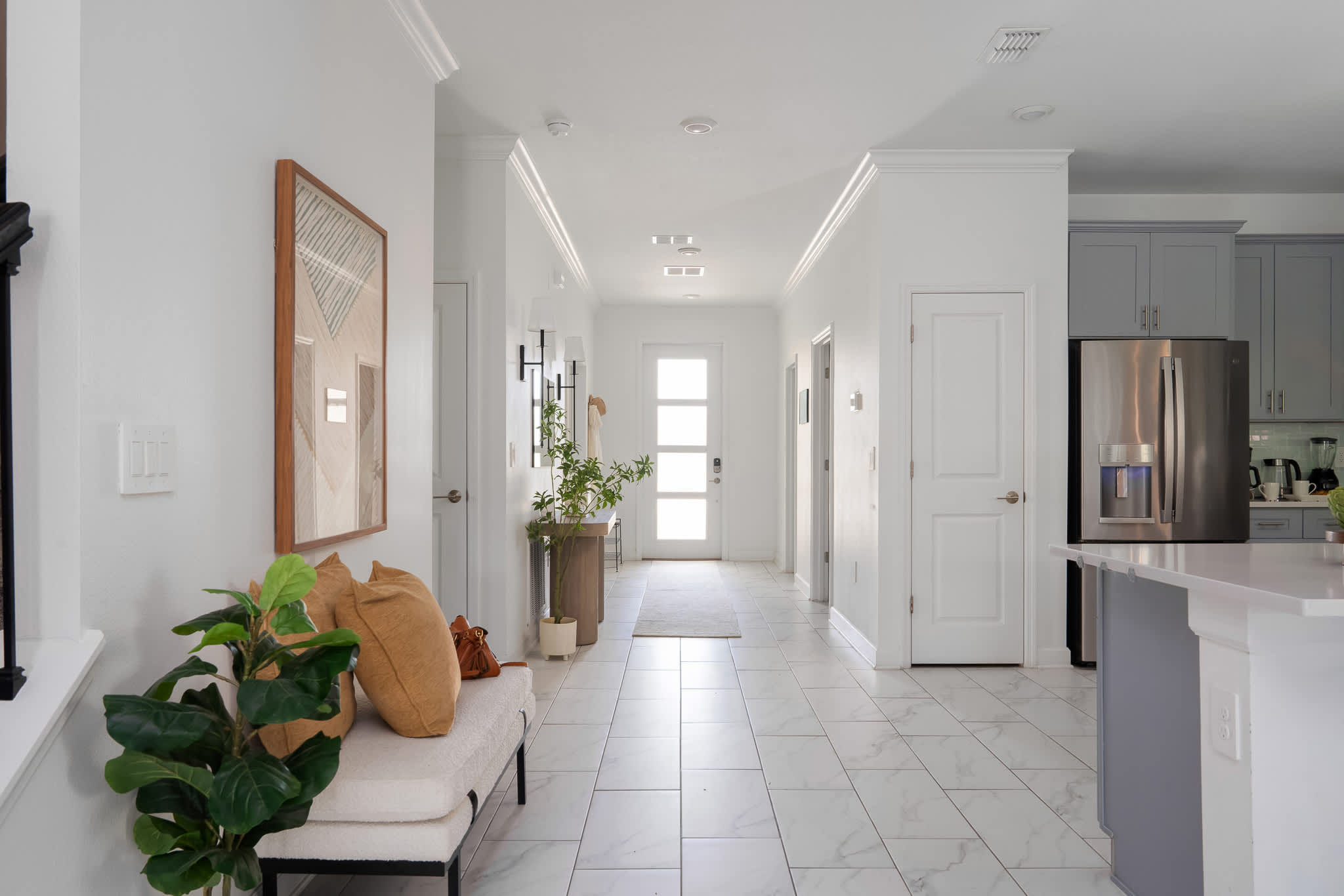 Bright and minimalist foyer with white tile flooring and walls, leading to a front door with frosted glass panels. A long bench with mustard yellow pillows and a large mirror decorates the space.