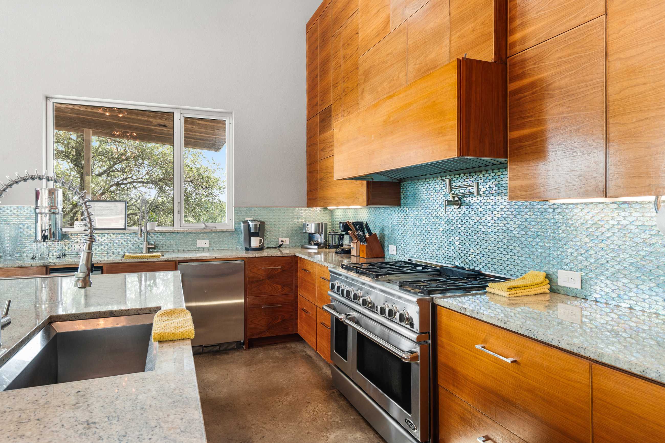 A detailed view of the kitchen's cooking area, showcasing a professional-grade stainless steel range and oven, with a light blue mosaic tile backsplash and granite countertops. Wooden cabinetry provides ample storage, and a large window offers natural light.
