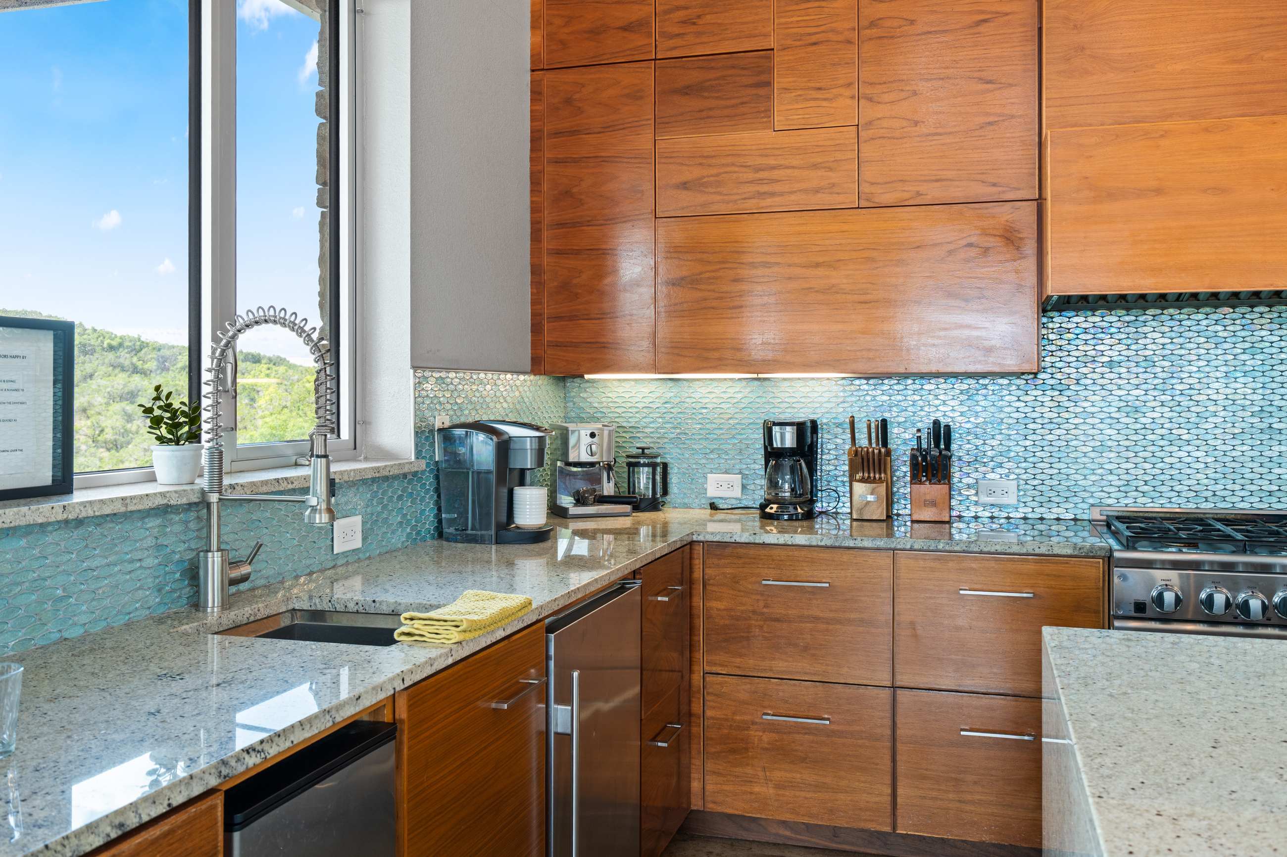 Section of a modern kitchen with a granite countertop, wooden drawers, and a blue mosaic tile backsplash, featuring a sink with a pull-down faucet and various kitchen appliances. A large window offers a bright outdoor view.