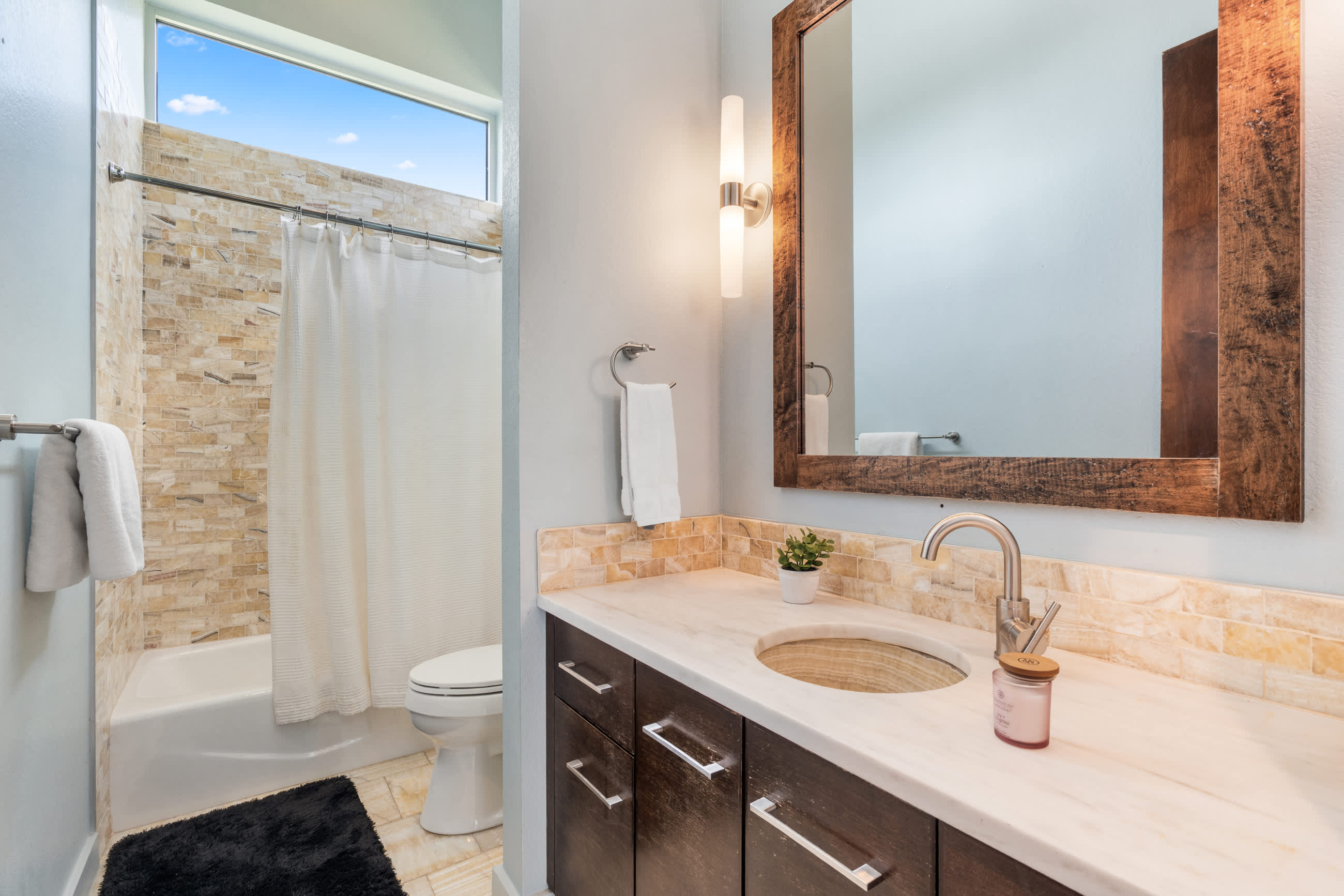 Modern bathroom with a single vanity featuring a light marble countertop, a wooden framed mirror, and a shower/tub combo with stacked stone tile walls and a small high window.