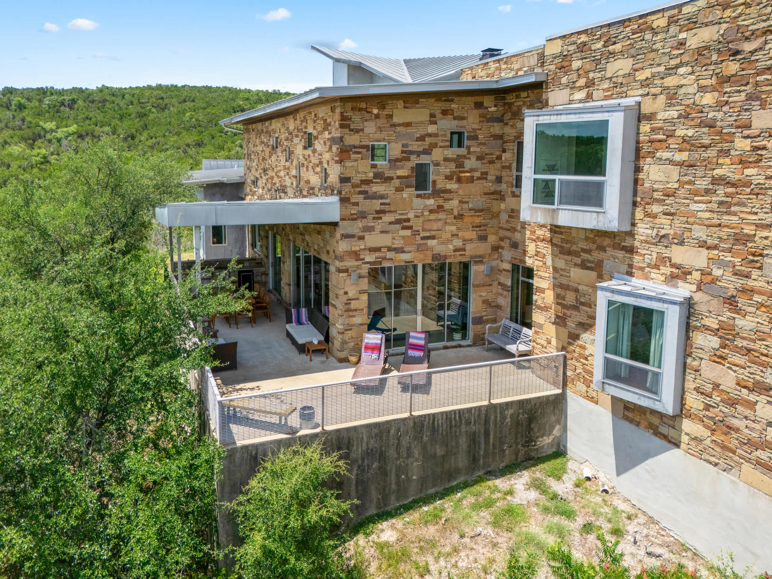An aerial perspective captures the multi-level modern house with stone and metal siding, featuring a large outdoor patio with lounge seating, overlooking a dense green landscape and distant hills.