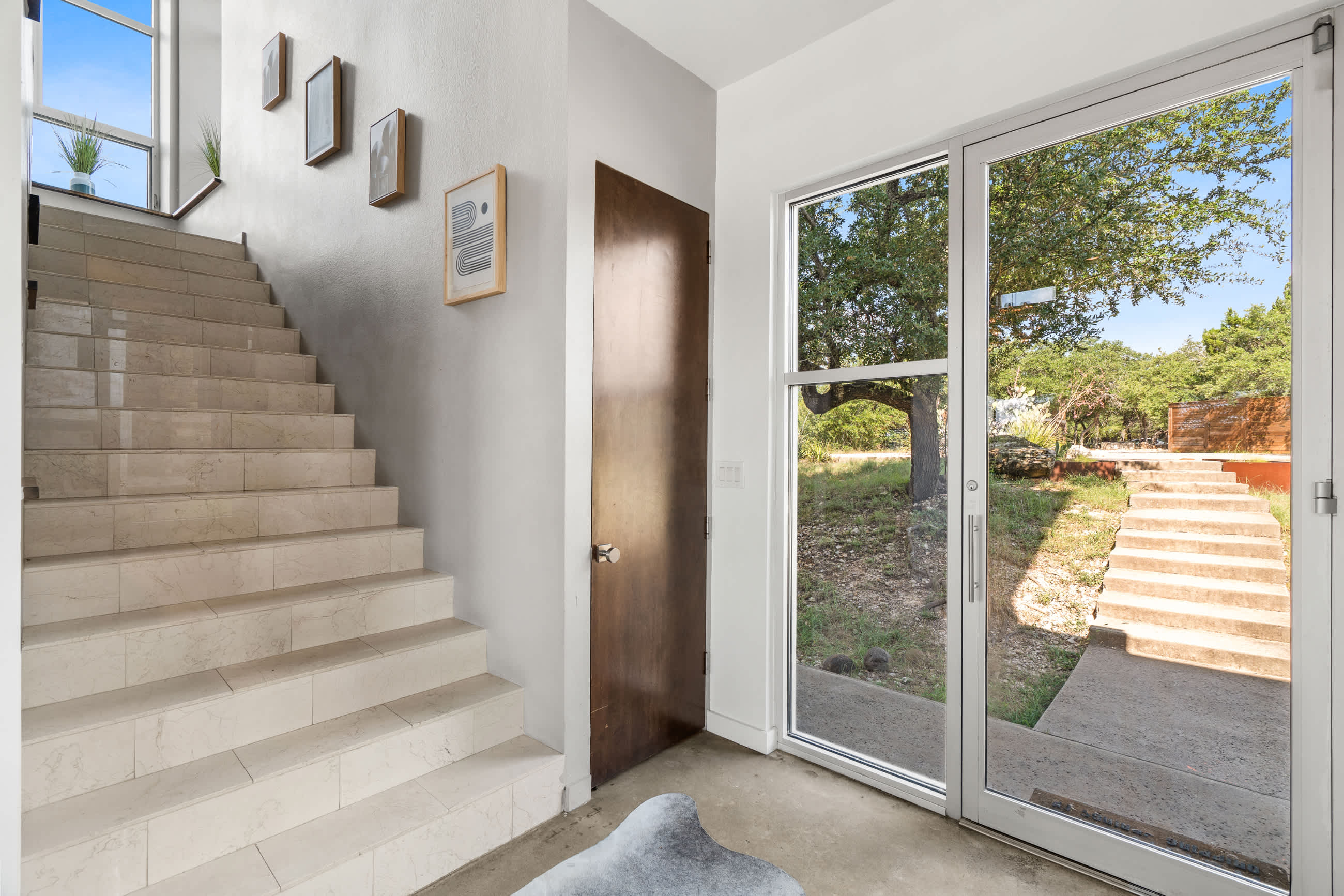 A modern entryway featuring a light-colored staircase with framed artwork on the wall, a dark wooden door, and large glass doors providing a view of outdoor stone steps and greenery. A gray cowhide rug is on the concrete floor.