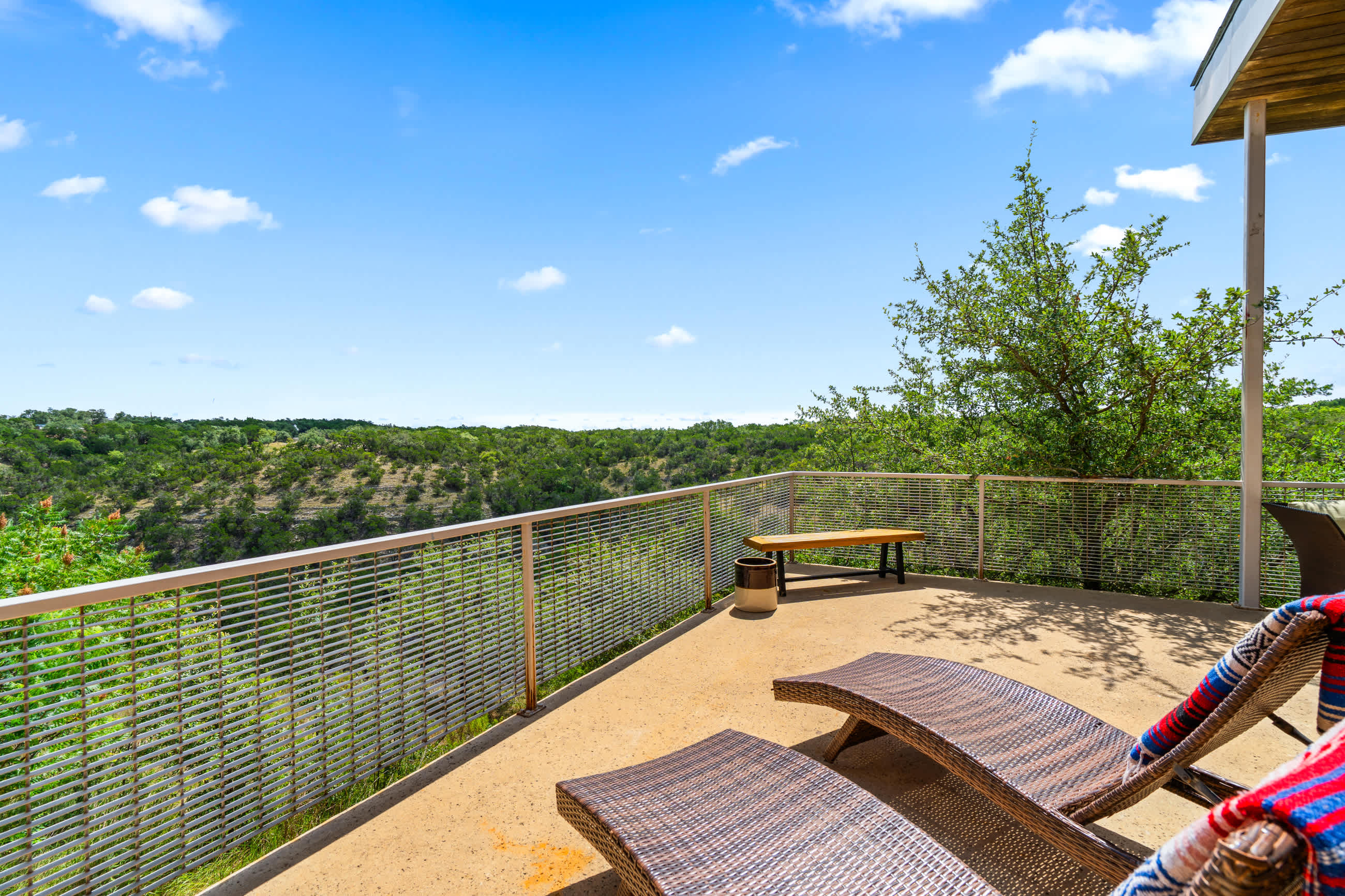 Two wicker lounge chairs with colorful blankets are placed on a concrete patio, facing a breathtaking panoramic view of rolling green hills under a bright blue sky, enclosed by a modern metal railing.