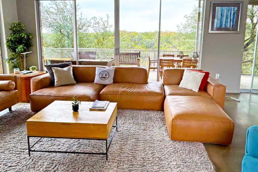 A cozy living area with a large brown leather sectional sofa arranged around a square wooden coffee table on a textured area rug. Floor-to-ceiling windows provide abundant natural light and views of the outdoor deck and greenery.