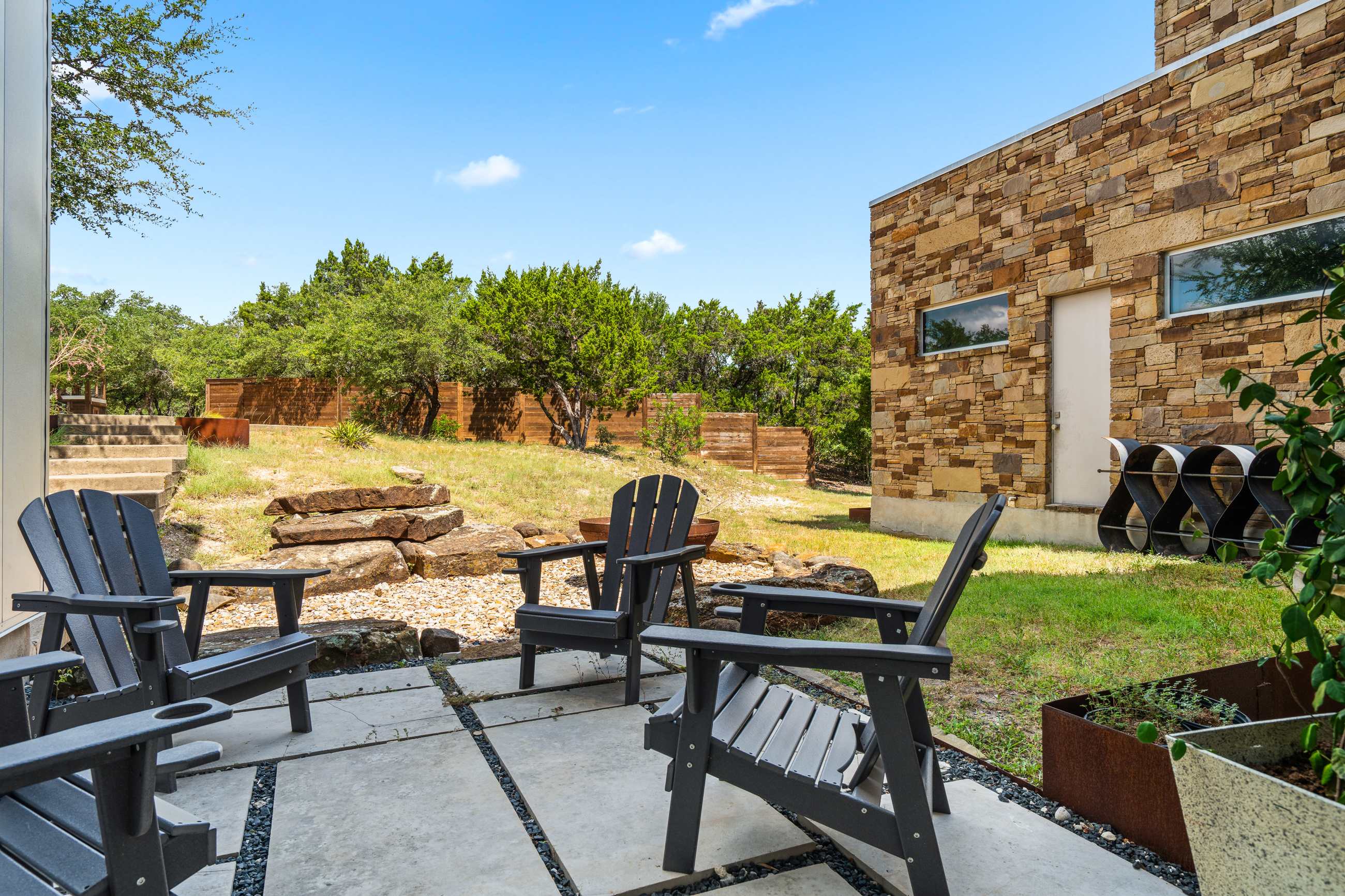 An inviting outdoor patio with gray Adirondack chairs on concrete pavers, overlooking a natural yard with gravel, large rocks, and lush trees, with the stone-clad house facade in the background.