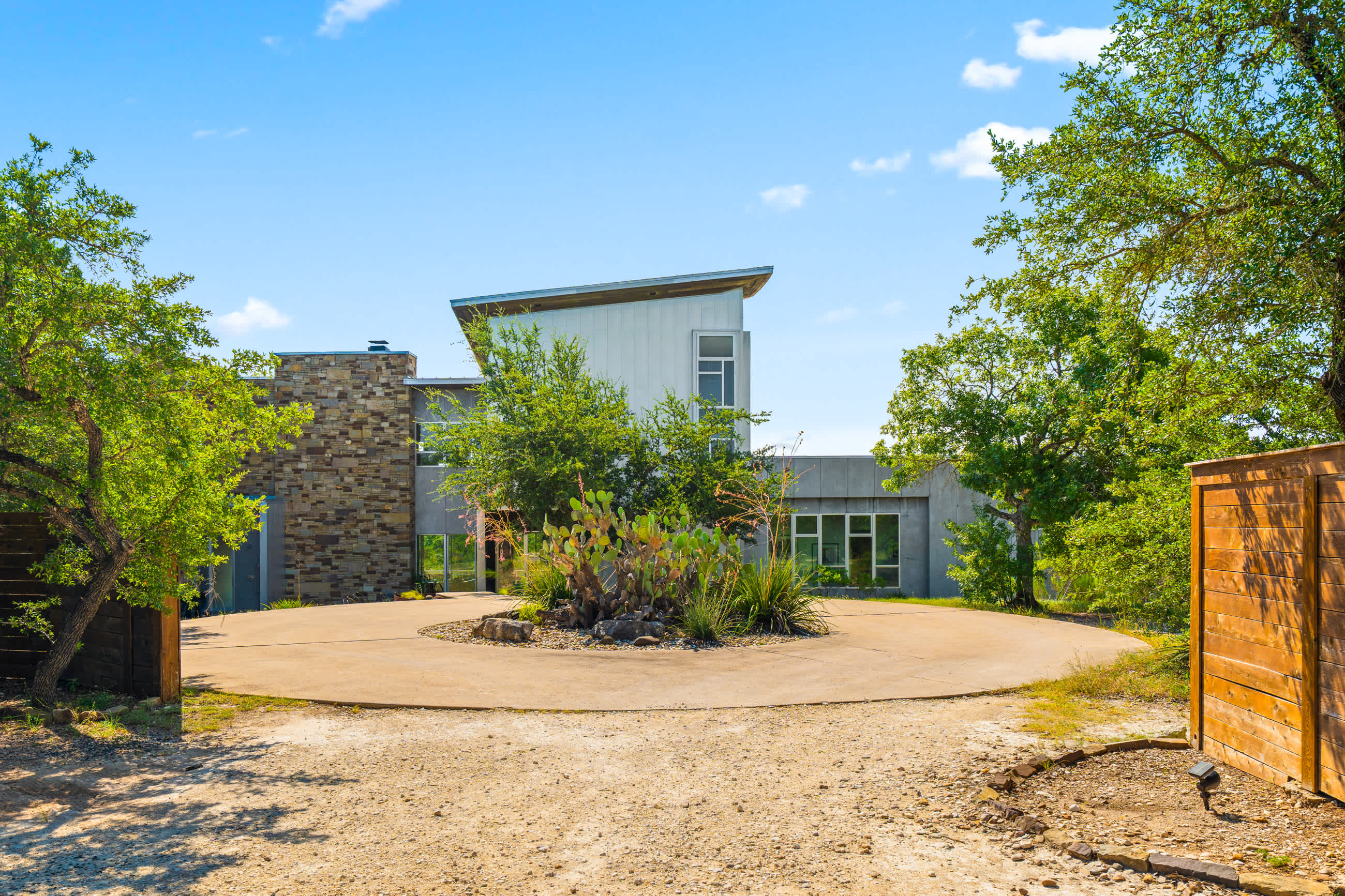 A contemporary house with a circular concrete driveway, featuring a mix of stone and light-colored siding, surrounded by mature trees and desert landscaping under a clear blue sky.