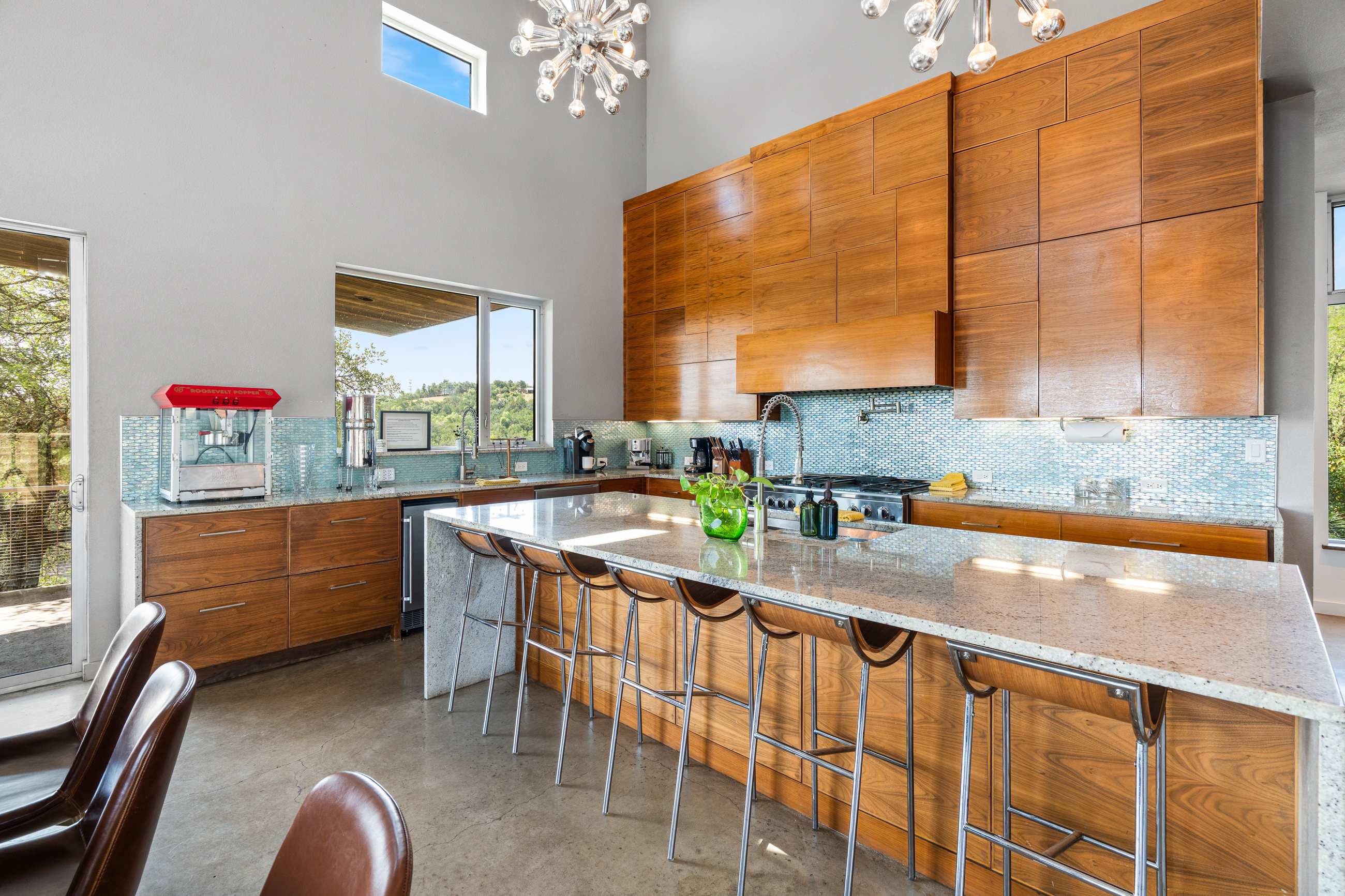 A bright kitchen with a large granite-topped island featuring bar seating and a sink, surrounded by sleek wooden cabinetry and light blue mosaic tile backsplash. A popcorn machine sits on the counter, and large windows overlook the outdoors.