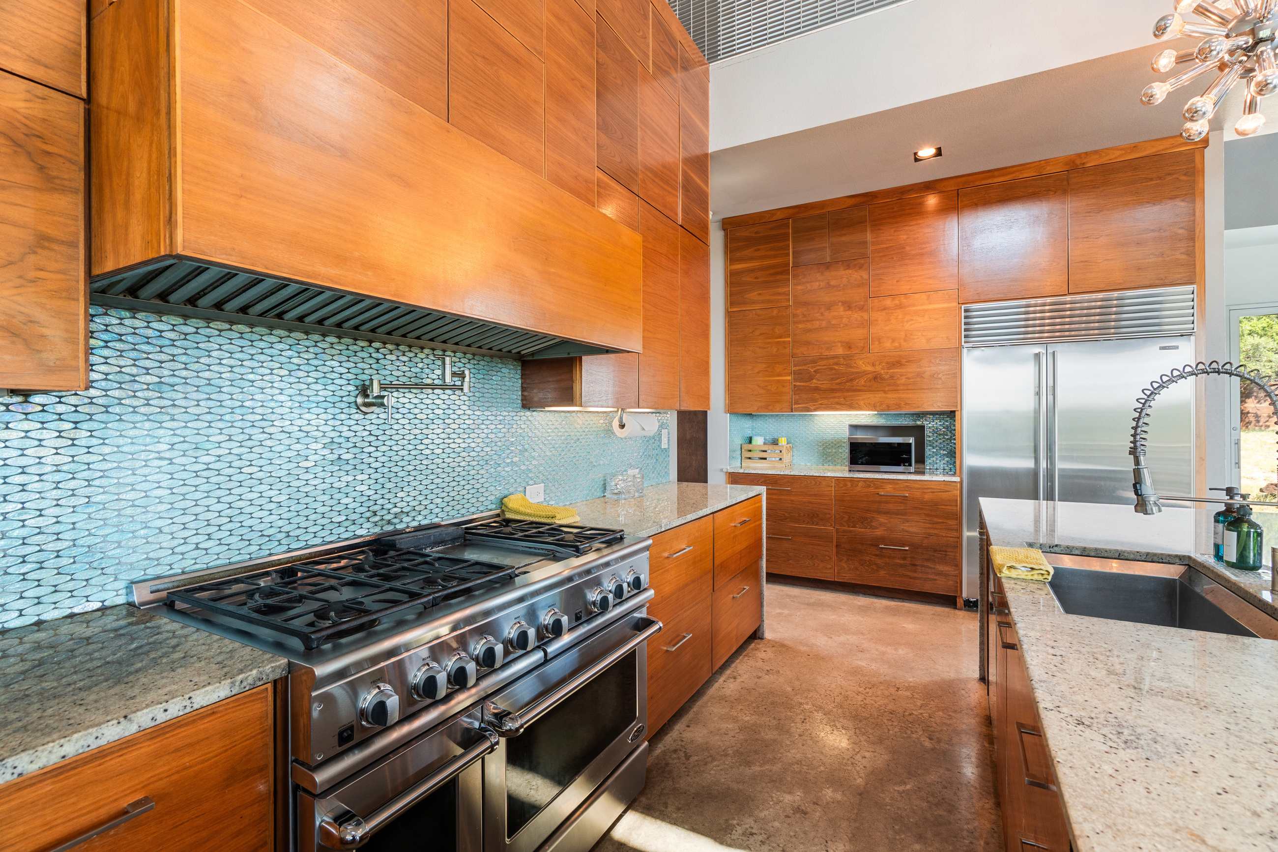 A close-up view of the kitchen's cooking area, featuring a large stainless steel range with multiple burners and double ovens, set against a light blue mosaic tile backsplash. Sleek wooden cabinetry extends to the high ceiling, with granite countertops.