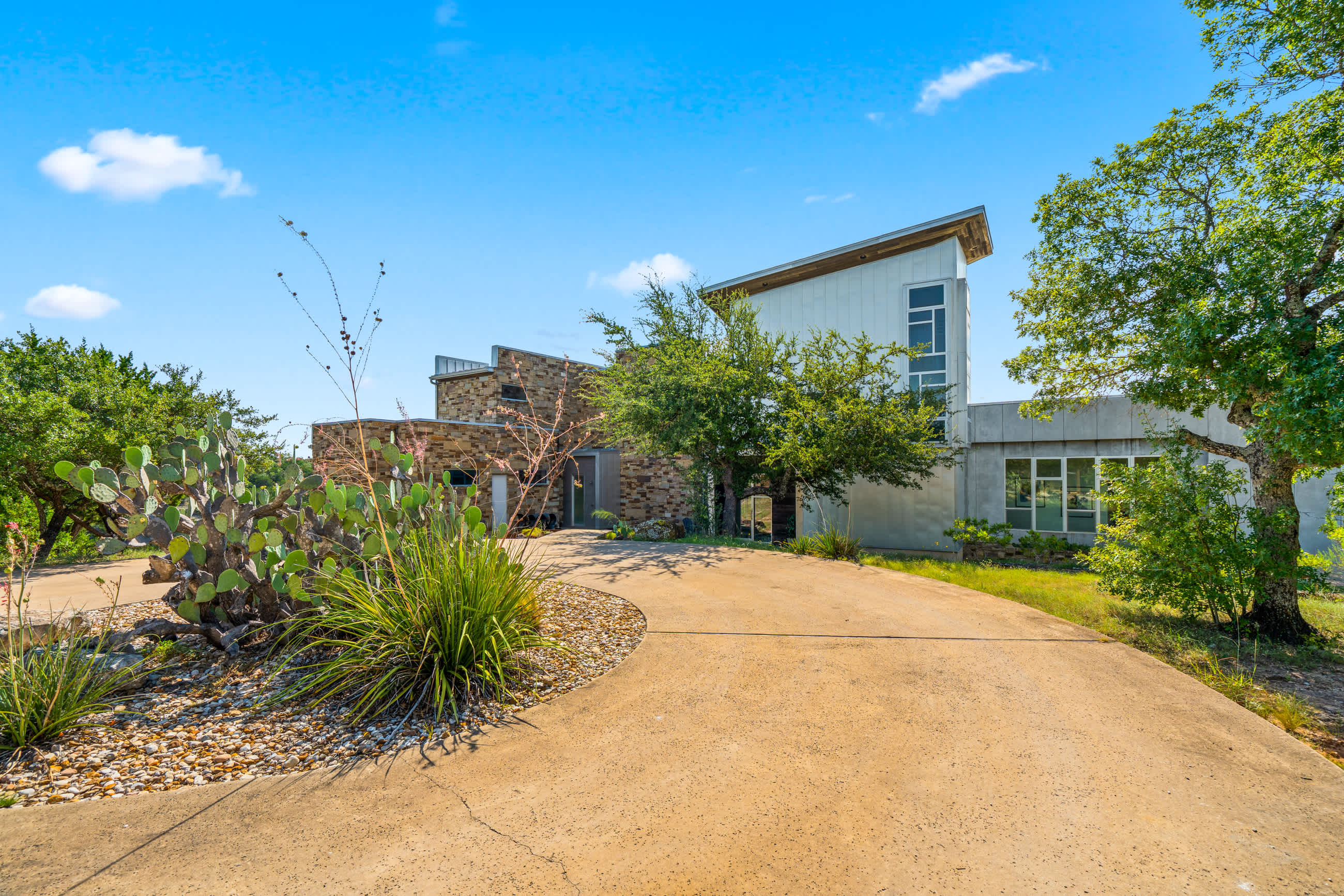An expansive concrete driveway winding towards a modern house with mixed materials like stone and light siding, set amidst natural landscaping with cacti, yucca plants, and mature trees.