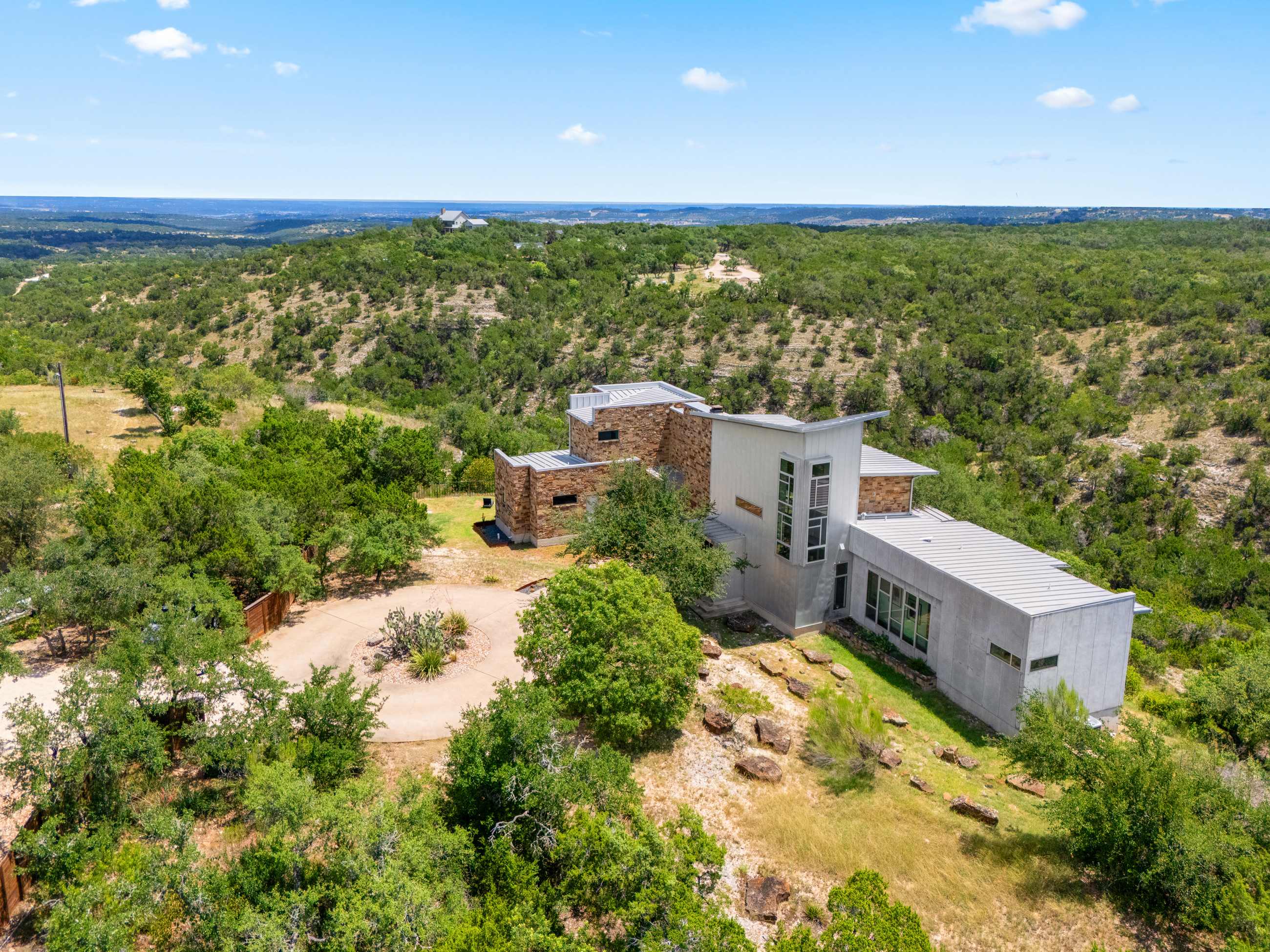 An aerial view showcases the modern house with stone and metal siding nestled amidst a vast, green, hilly landscape under a clear blue sky, with a winding driveway and sparse trees.