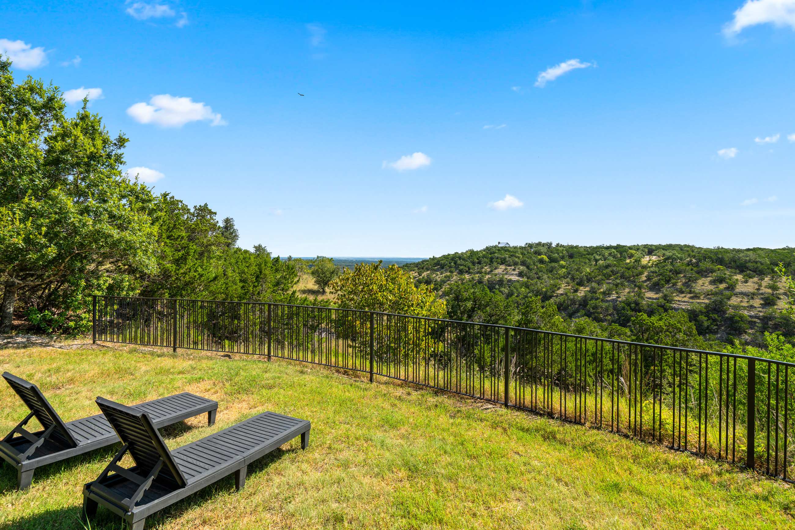 Expansive grassy yard with two dark lounge chairs positioned to enjoy panoramic views of a sprawling, tree-filled valley and distant hills under a bright blue sky. A black metal fence lines the edge of the property.