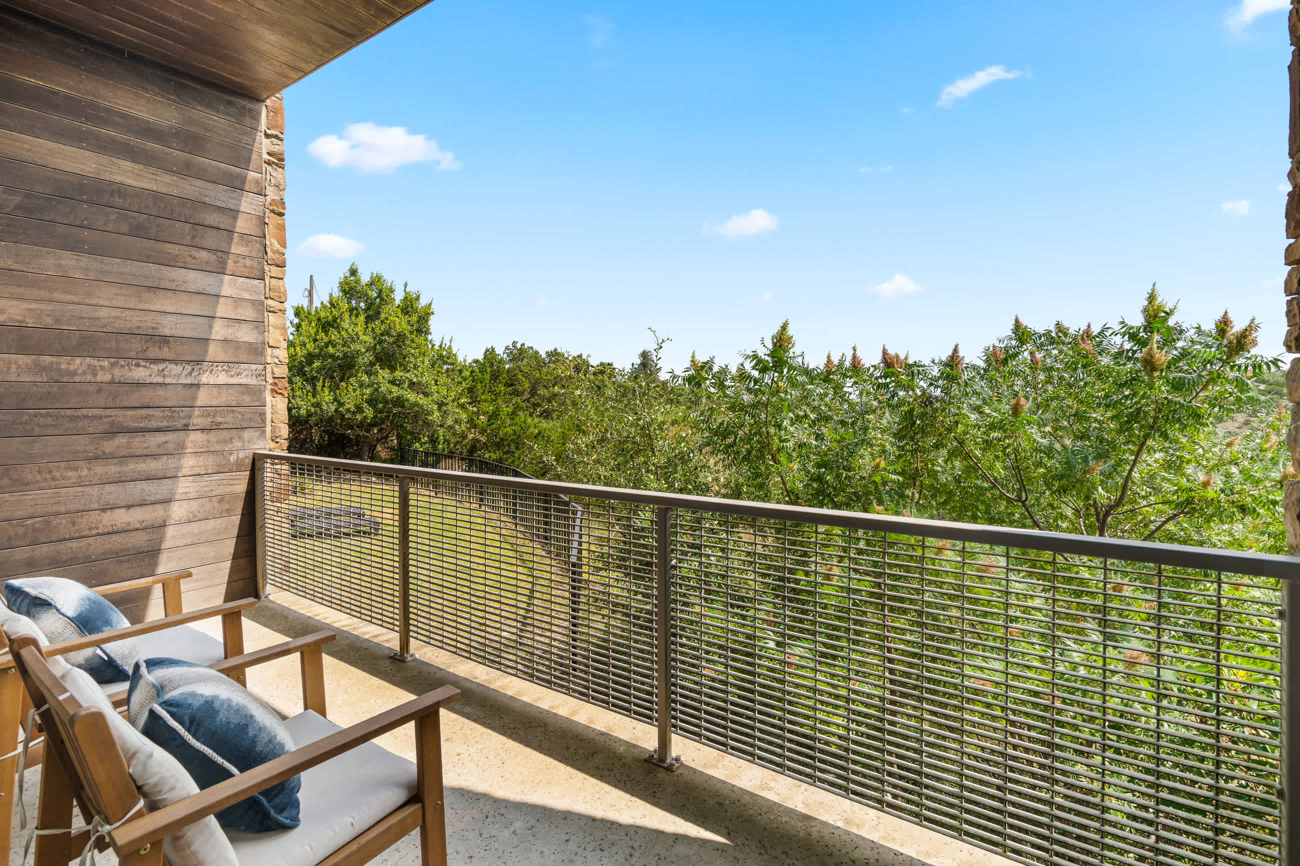 A private balcony features two wooden chairs with light cushions and blue throw pillows, set against a modern wood-paneled wall and a stone pillar. The metal railing overlooks a vibrant green landscape.