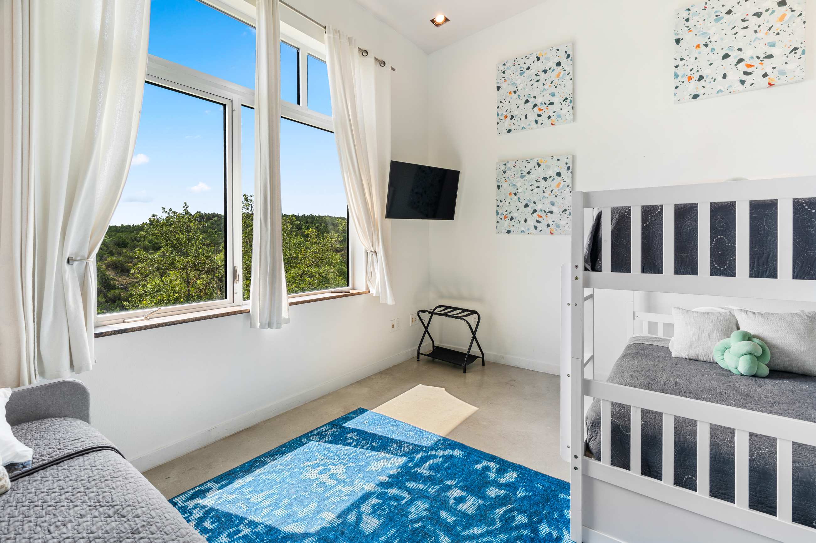 Bright bedroom with white walls, a white bunk bed, a gray daybed, and a vibrant blue rug, featuring large windows with scenic views of lush green hills. A wall-mounted TV and luggage rack are also present.